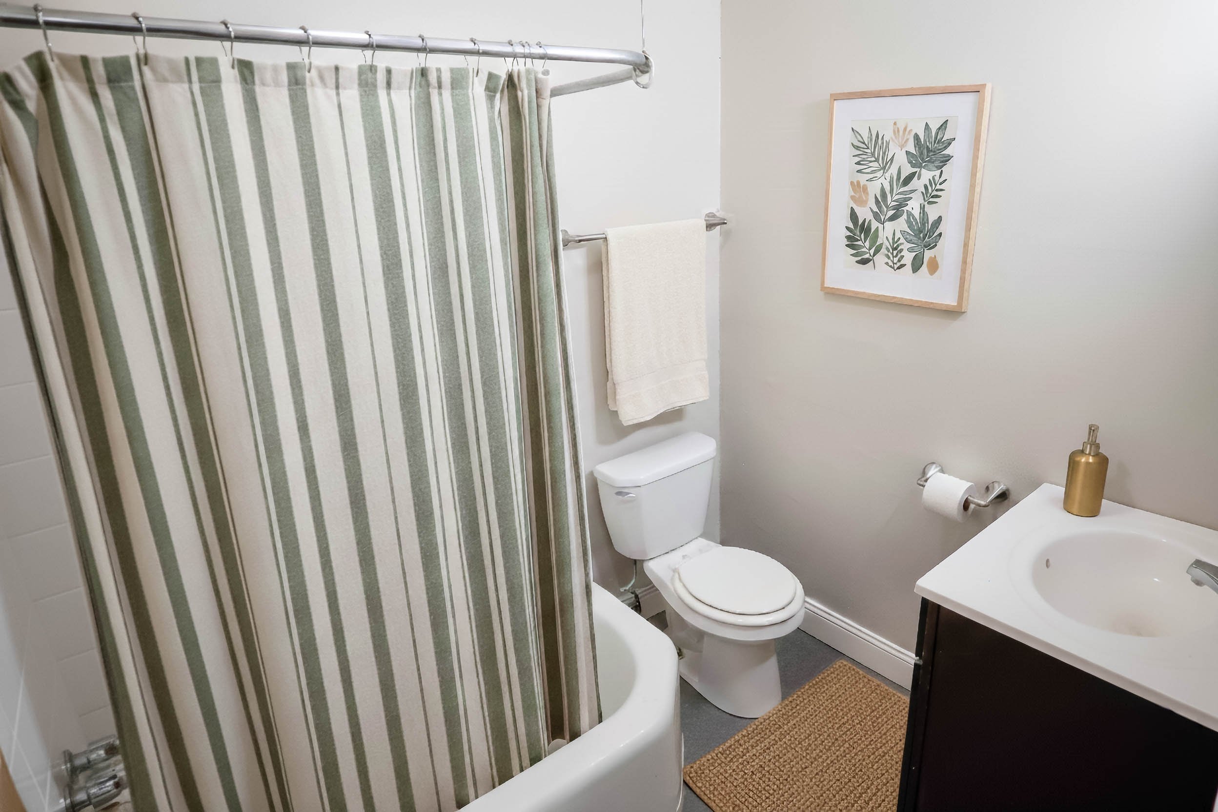 A small bathroom with a striped shower curtain, a toilet, a framed botanical print on the wall, a towel hanging above the toilet, a small sink with a soap dispenser, and a woven floor mat.