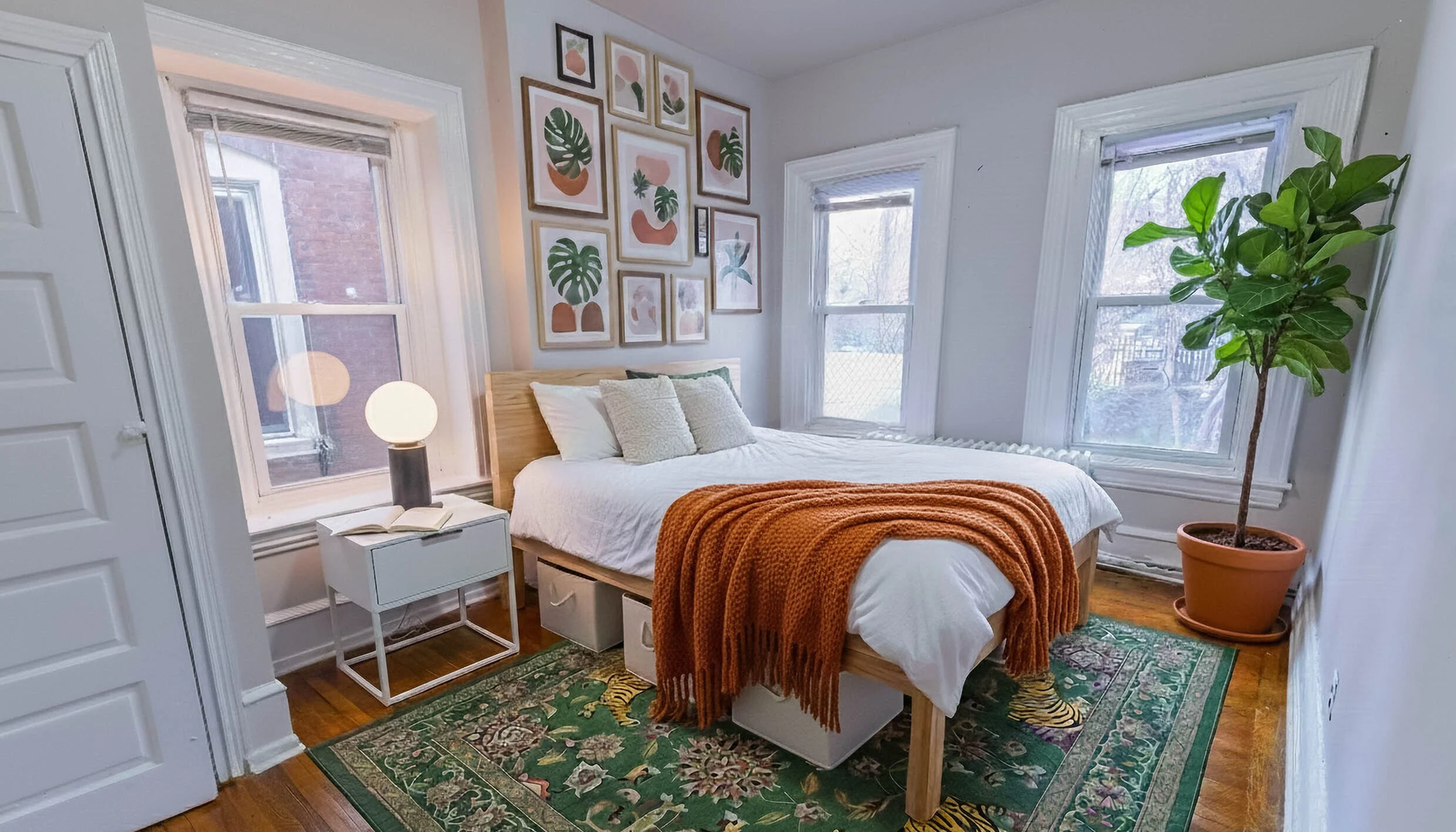 A cozy bedroom with a bed, side table, and decorative elements. The bed has white bedding, three pillows, and an orange throw blanket. A white nightstand with an open book and a lamp are next to the bed. The wall features a gallery of framed botanica