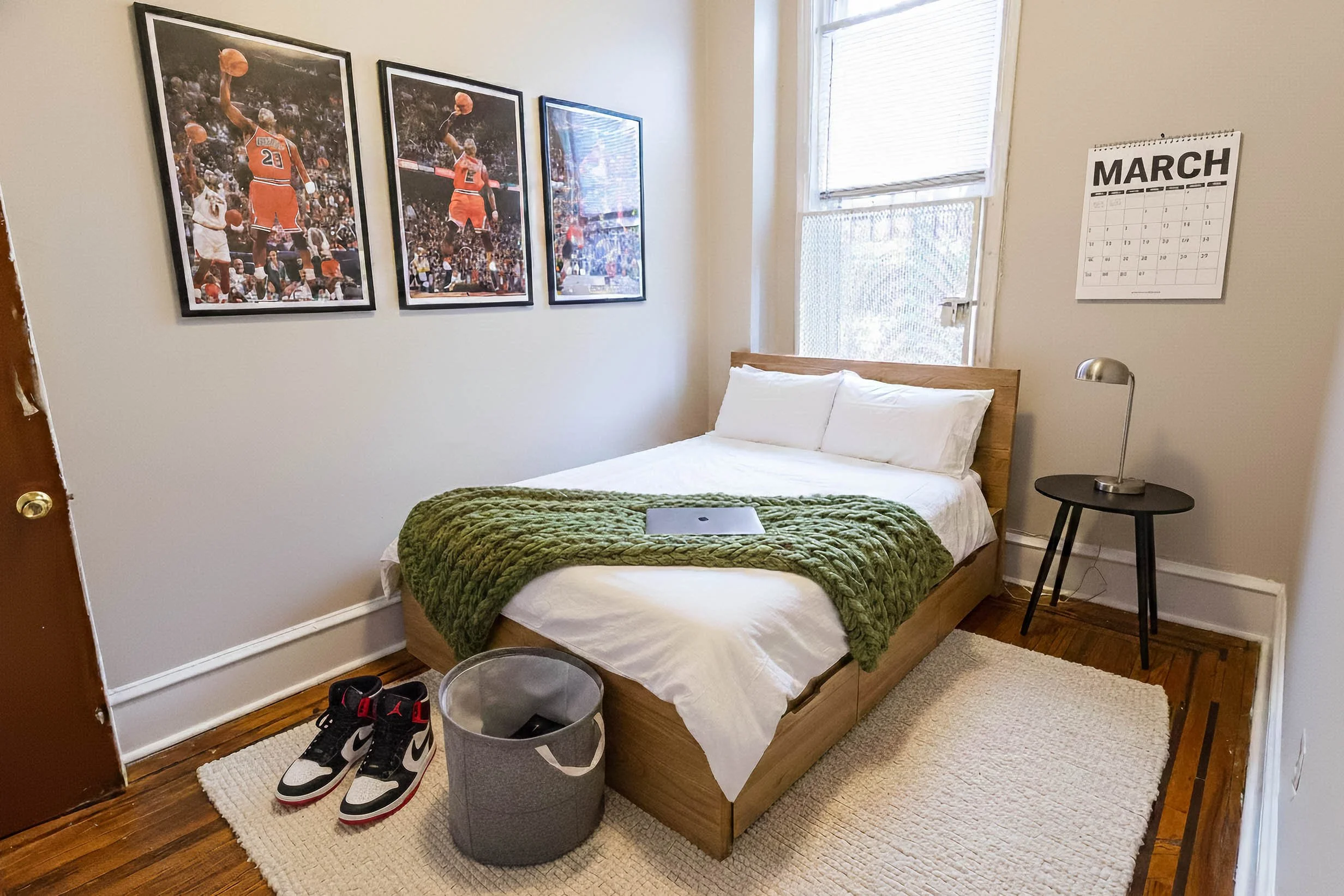 A bedroom with a wooden bed frame, white bedding, and a green throw blanket. A laptop is on the bed. On the left, there are sneakers on a white rug, with a gray laundry basket next to them. To the right, a small black side table with a silver lamp. B
