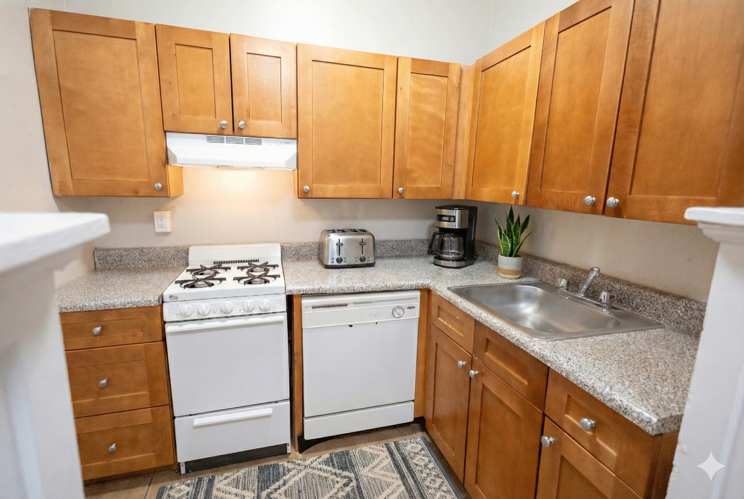 Small kitchen with wooden cabinets, white stove, dishwasher, coffee maker, toaster, and a potted plant on granite countertops.