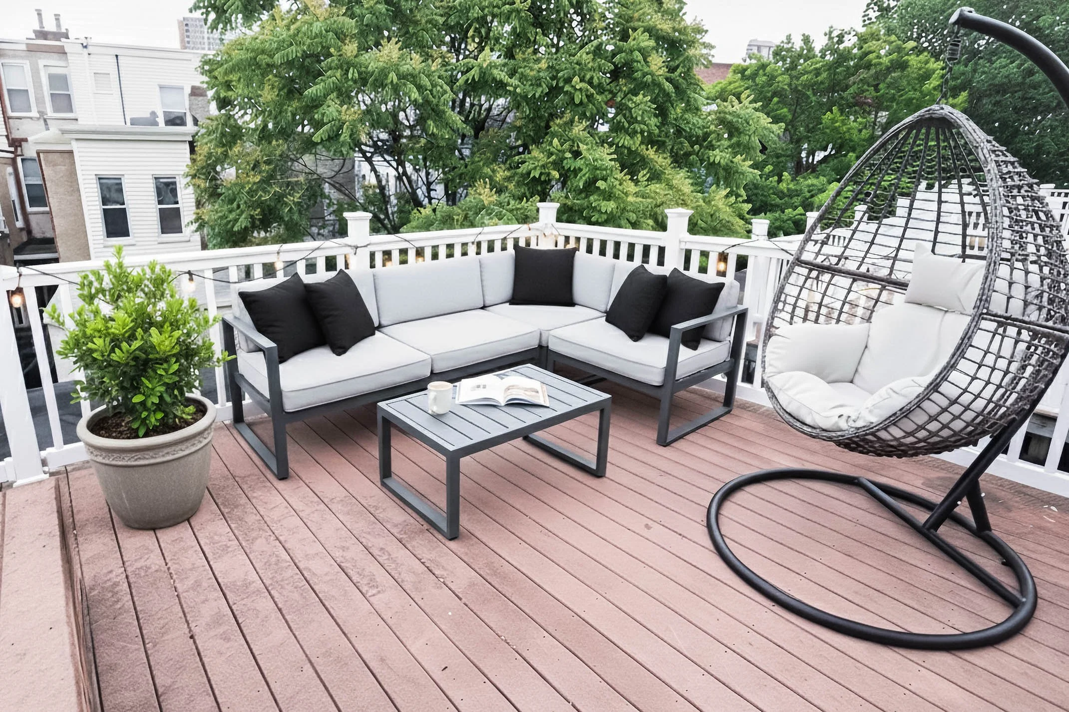 A rooftop patio with white outdoor sectional sofa with black cushions, a gray coffee table with a book and mug, a potted plant, and a hanging egg chair with white cushions. There are string lights and green trees in the background.