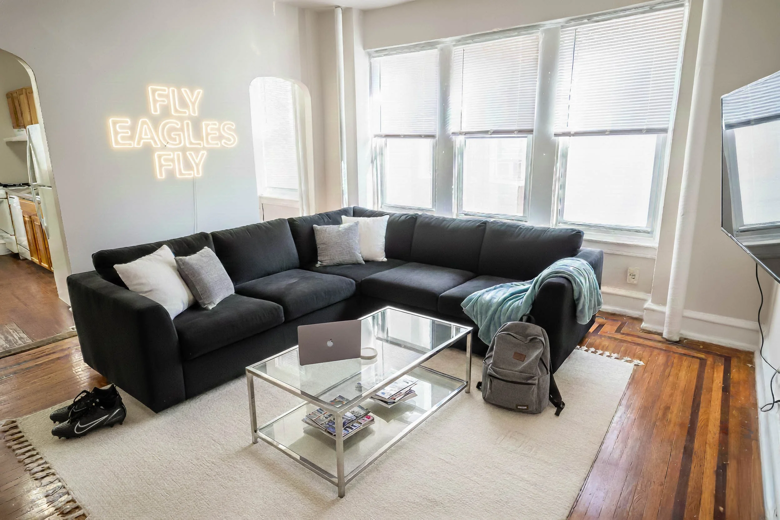 Living room with black sectional sofa, pillows, glass coffee table with magazines, a laptop, a backpack, a blanket, a TV mounted on the wall, large windows with blinds, and neon sign on the wall that says 'Fly Eagles Fly'