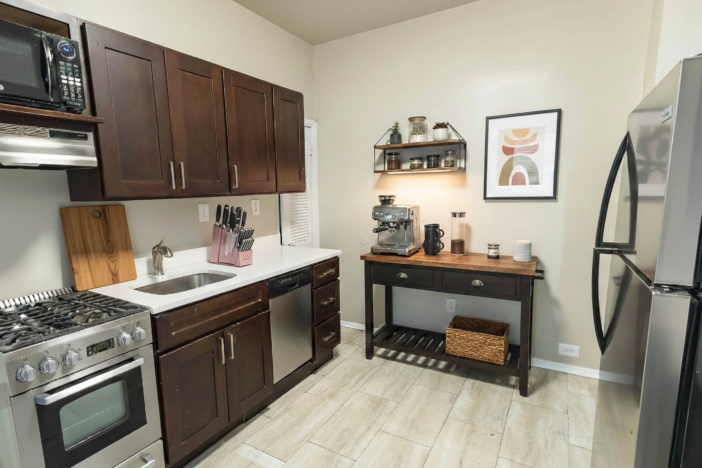 Kitchen with dark wood cabinets, stainless steel stove and dishwasher, white countertop, and coffee brewing station with decor on a wooden table, framed abstract art on the wall, and a black shelf with jars.