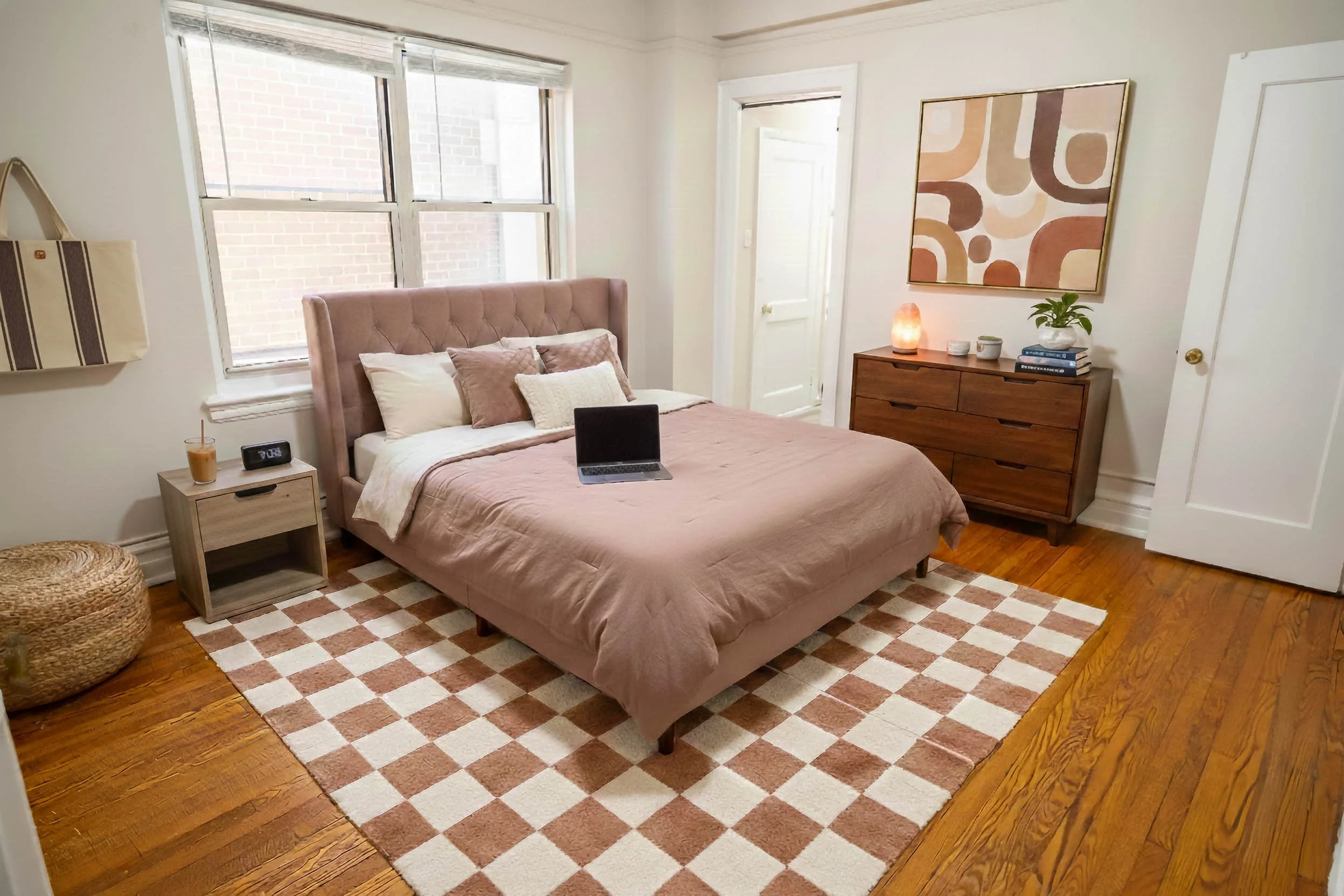A bedroom with a pink upholstered bed, a checkered rug, wooden furniture, and a laptop on the bed.