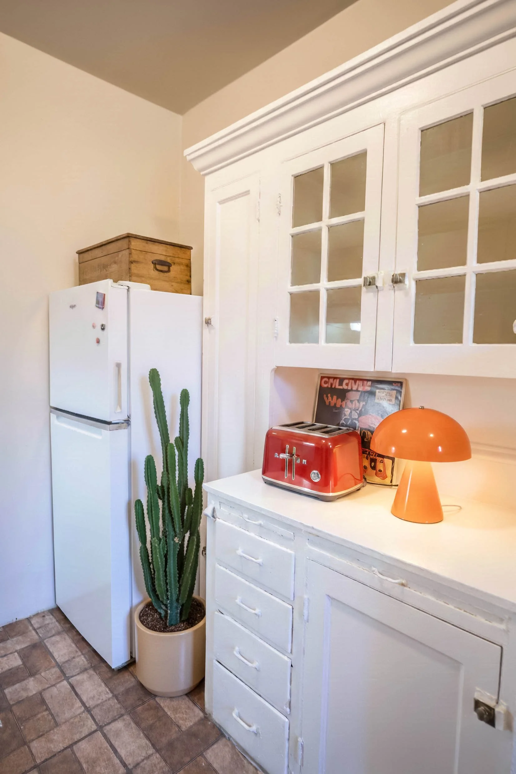 A kitchen corner with a white refrigerator, a tall cactus plant in a beige pot, vintage wooden box, white cabinet with drawers, red toaster, orange table lamp, and a vinyl record.
