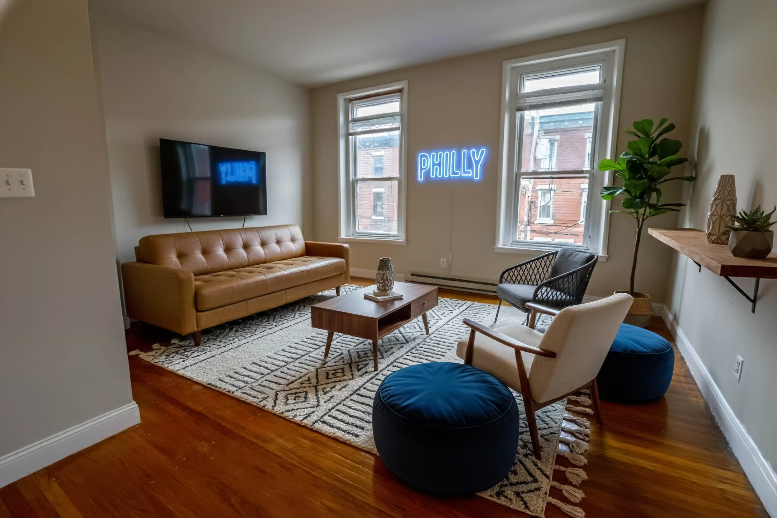 Living room with a beige sofa, a wooden coffee table, two armchairs, a TV mounted on the wall, a blue neon sign that says 'PHILLY', a large potted plant, and decorative items on a floating shelf.