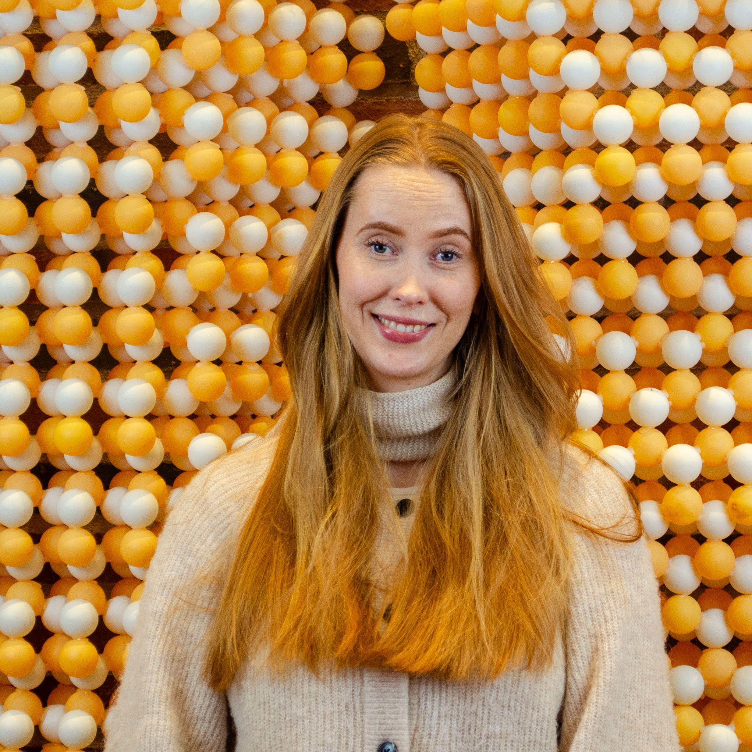 A woman with long red hair smiling at the camera in front of a background of orange and white plastic balls.