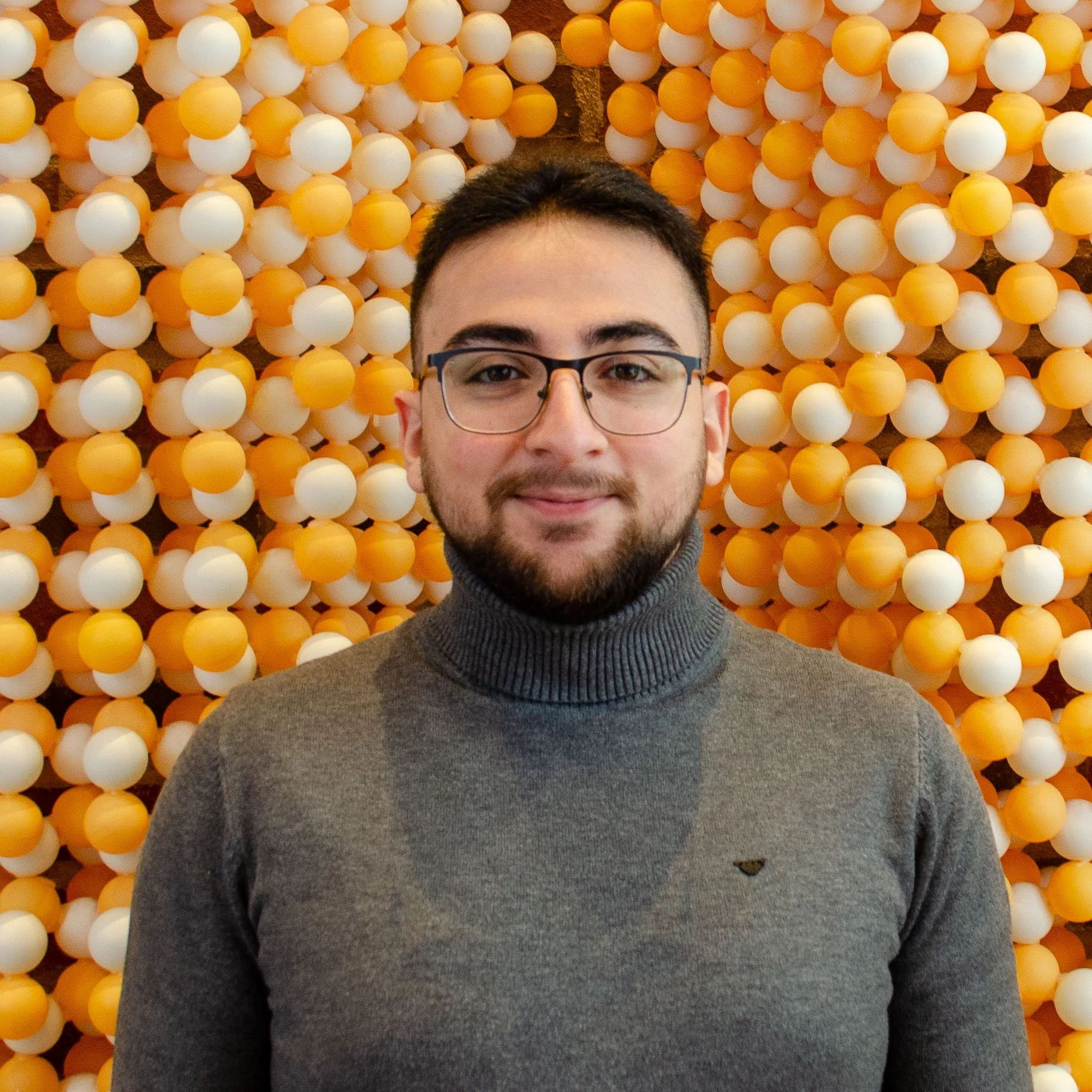 A young man with glasses and a beard, wearing a gray turtleneck sweater, standing in front of a background of orange and white round decorative balls.