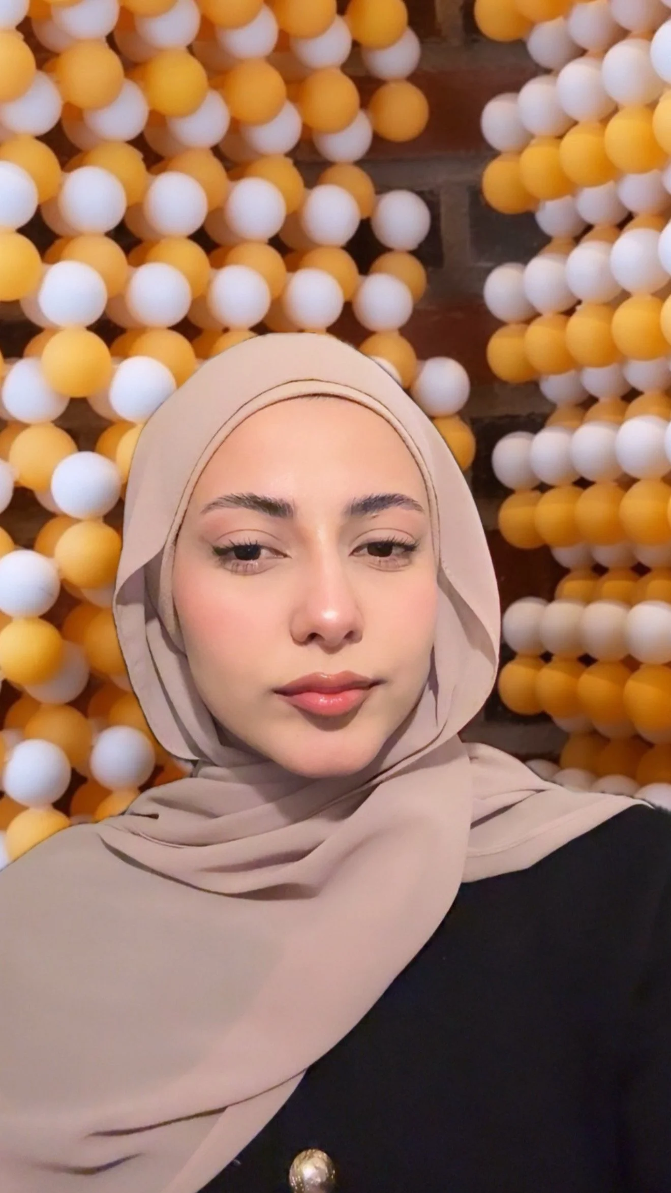 A woman in a beige hijab taking a selfie in front of a wall decorated with yellow and white spherical ornaments.
