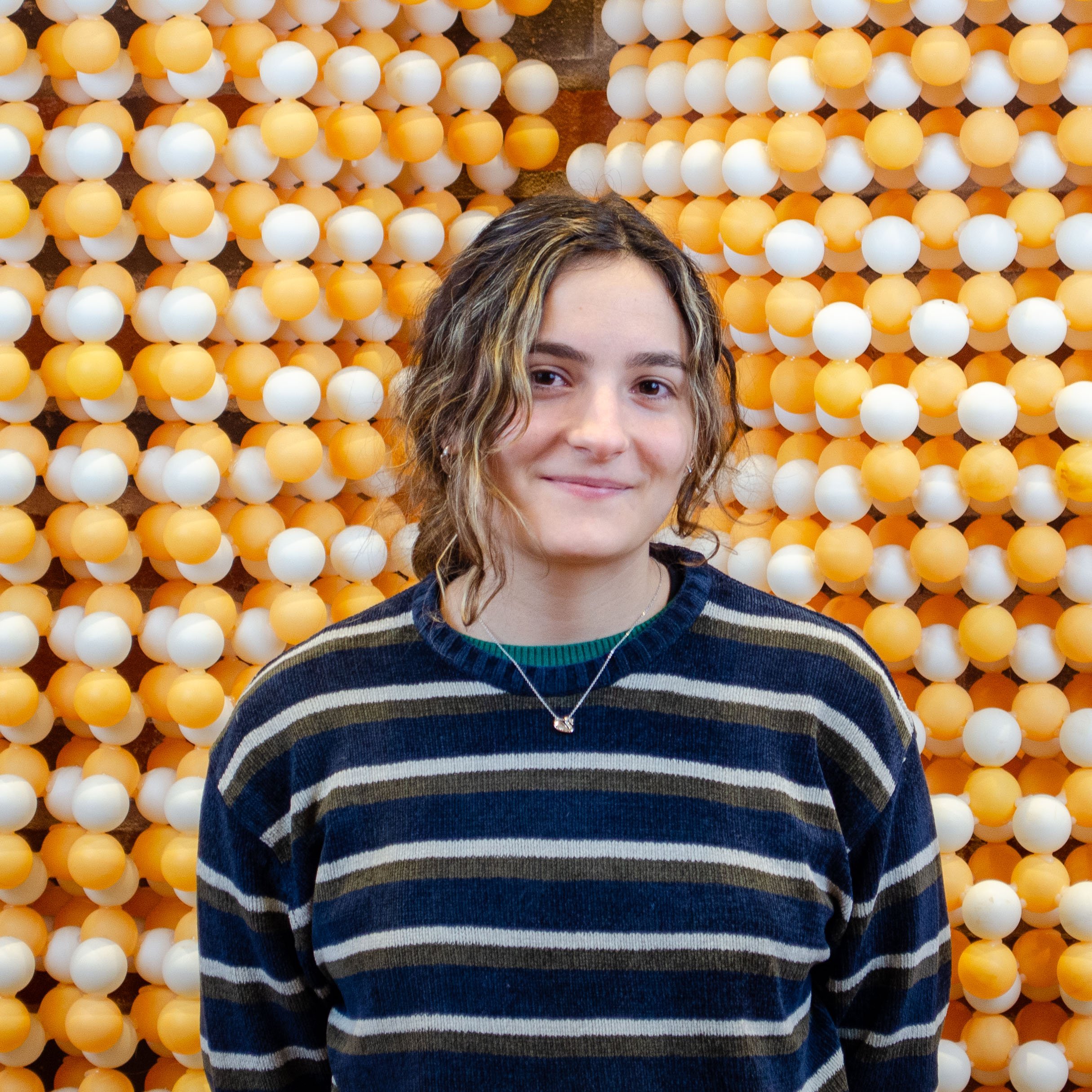 A young woman with wavy light brown hair and a striped sweater standing in front of a wall of yellow and white plastic balls.