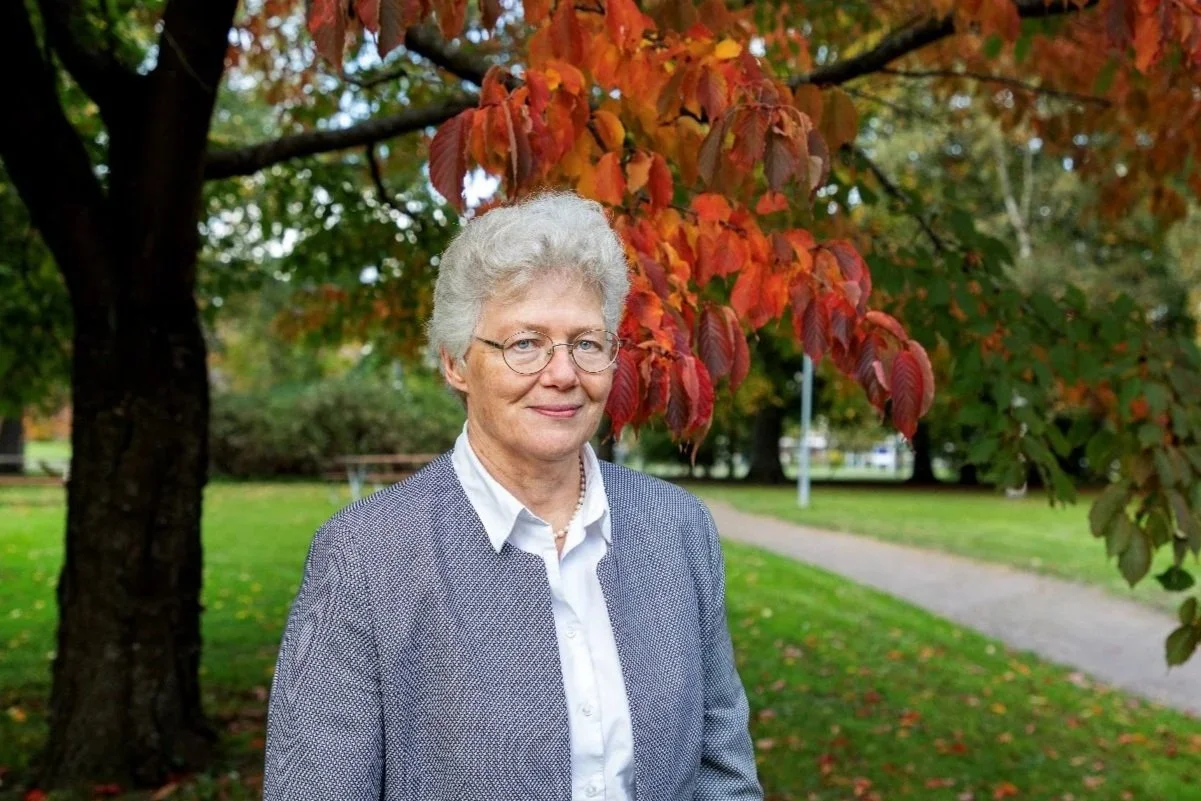An older woman with glasses and gray curly hair standing outdoors in a park during fall, wearing a gray blazer over a white shirt, with autumn red and orange leaves on a tree behind her.