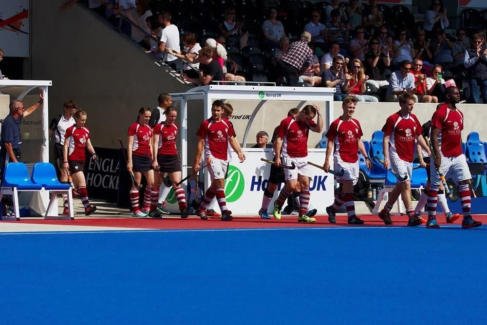 Field hockey players in maroon kit walking onto the field from the side lines, with spectators seated in the background.