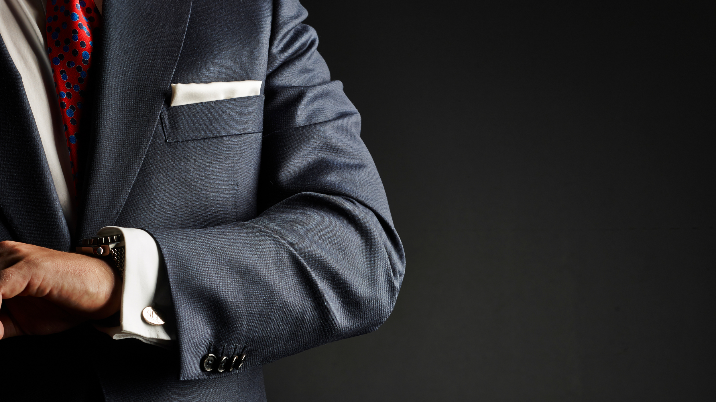 Close-up of a man in a dark gray suit with a white shirt, red polka dot tie, and a white pocket square, adjusting his watch against a black background.