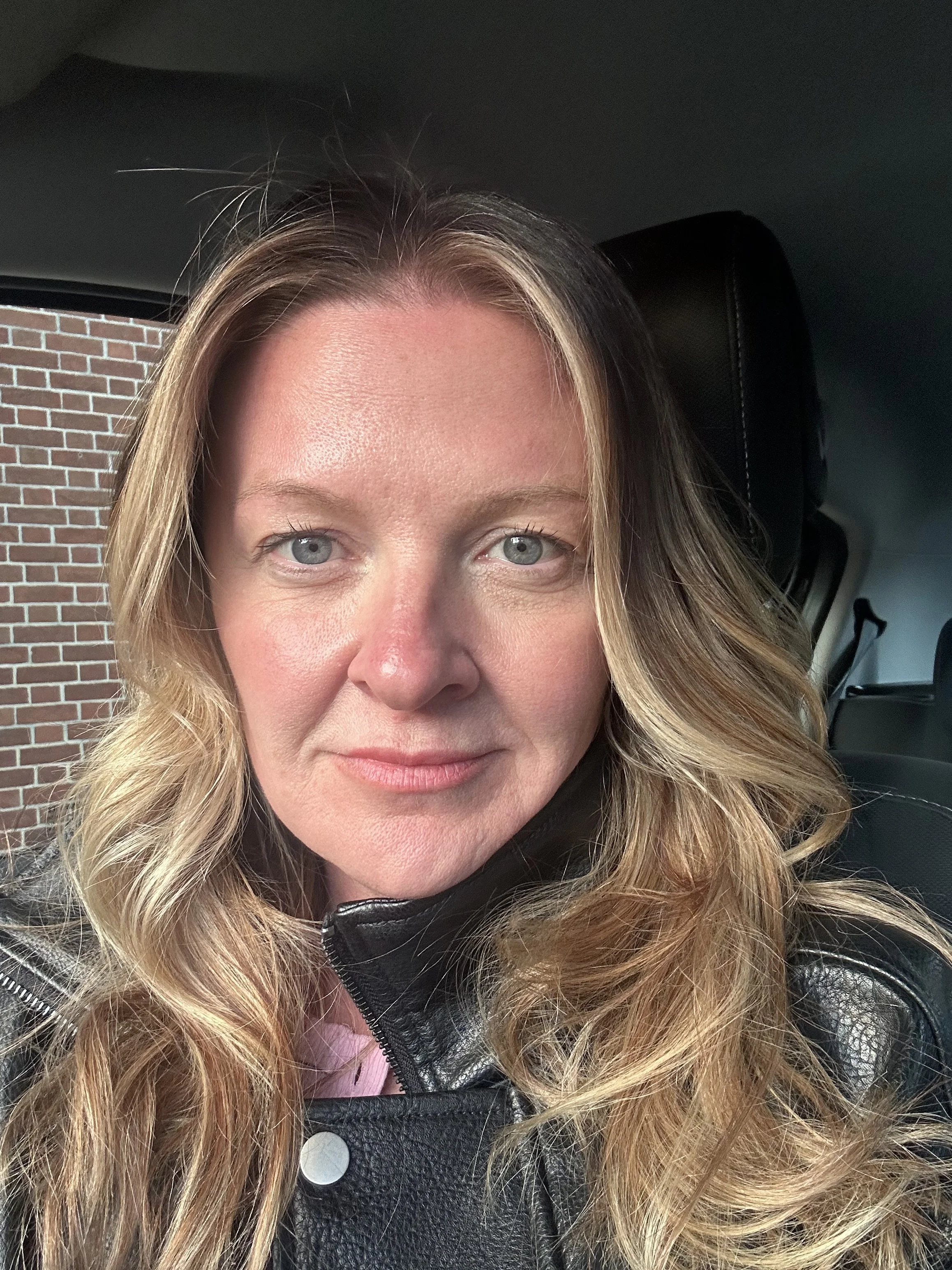 Close-up of a woman's face with blonde wavy hair and blue eyes, wearing a black leather jacket, sitting inside a vehicle with a brick wall visible in the background.