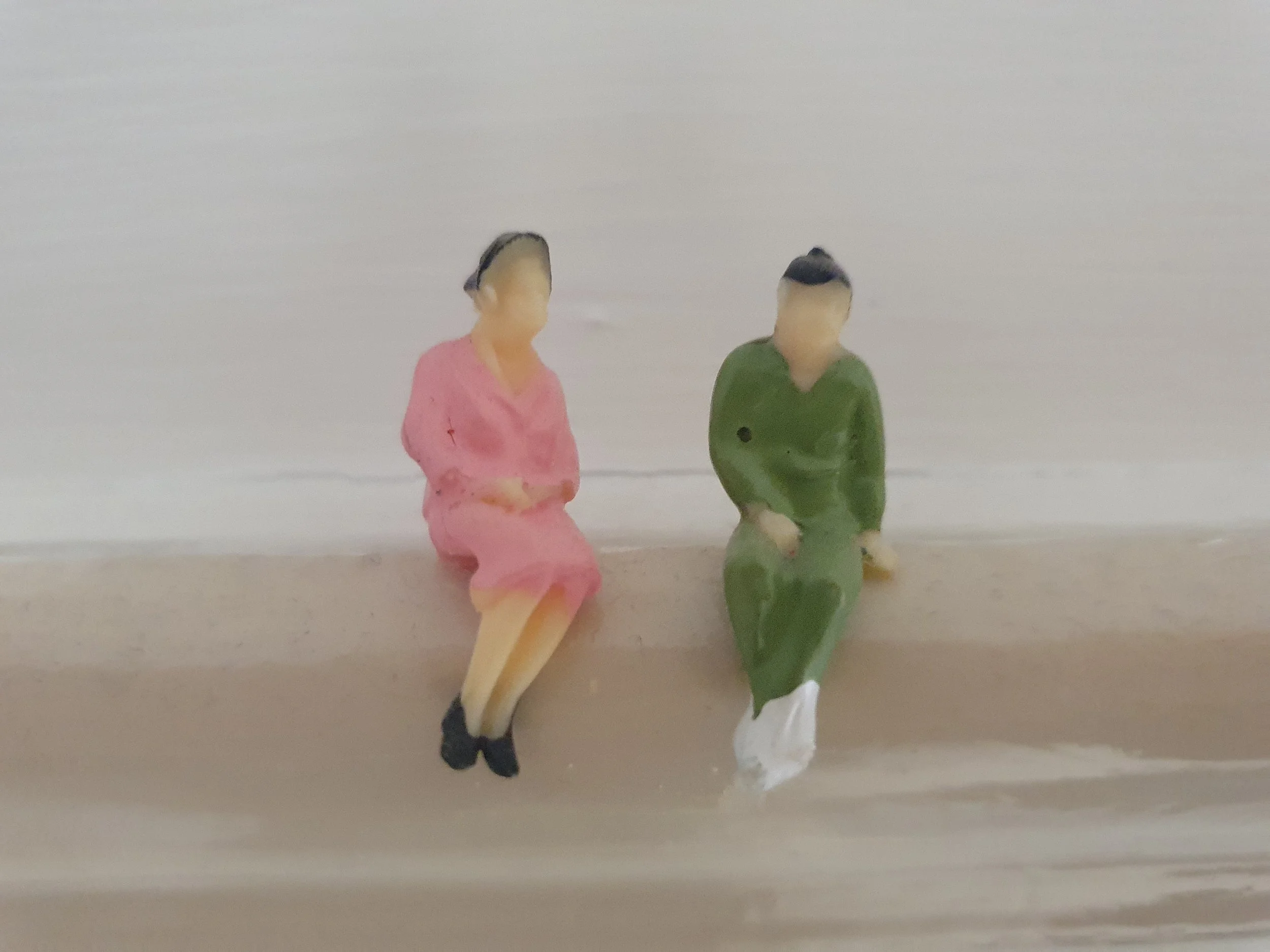 Two small figurines facing a wall on a shelf, one in a pink outfit and the other in a dark outfit.