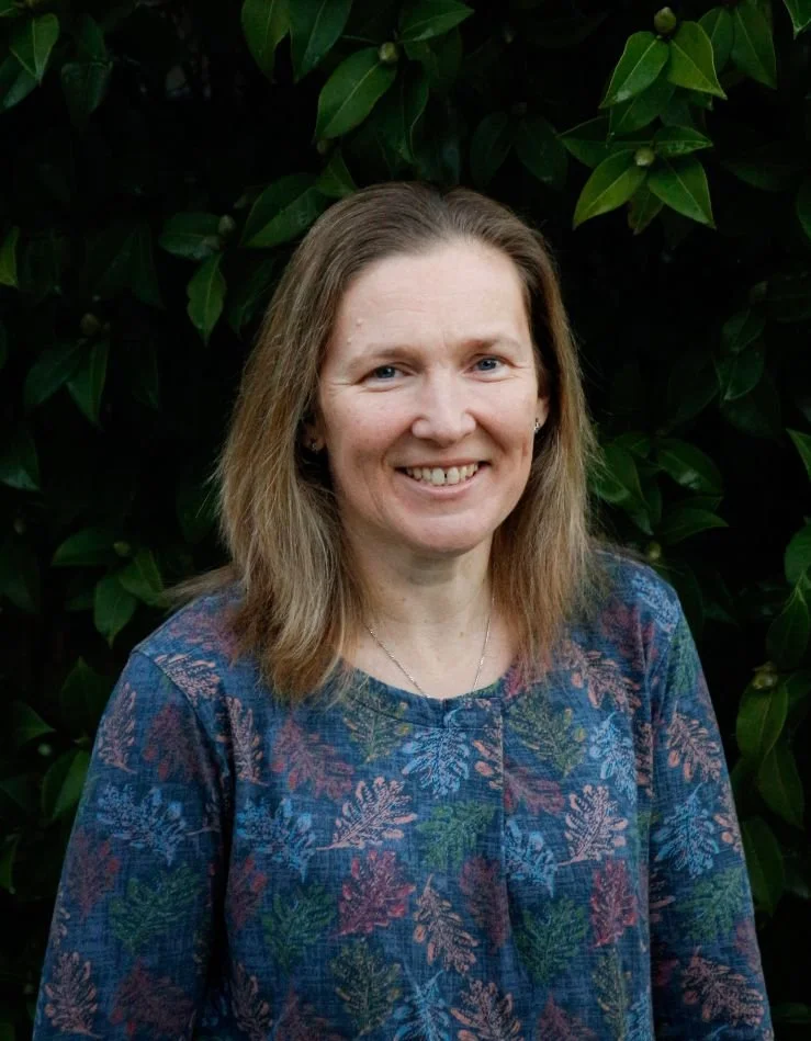 A woman with shoulder-length light brown hair smiling, standing outdoors in front of dark green leafy bushes, wearing a blue patterned blouse.