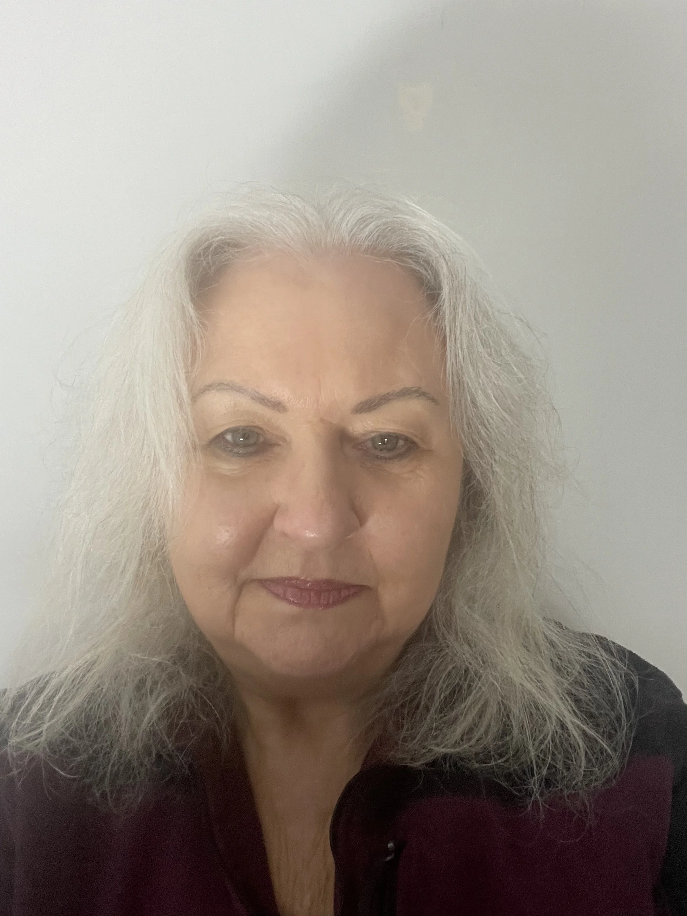 A close-up of an woman with long, grey hair and light skin, wearing a dark-colored top, against a plain light background.