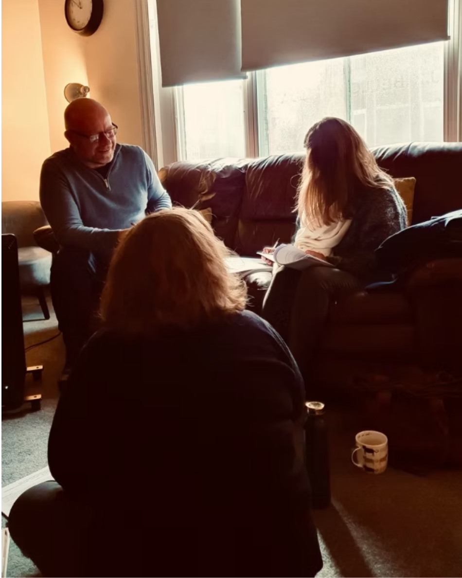 Three students sitting in a living room near a window, engaged in conversation. The man is smiling, and two women are reading a notebook and a book. A mug is on the floor in front of the woman with her back to the camera.