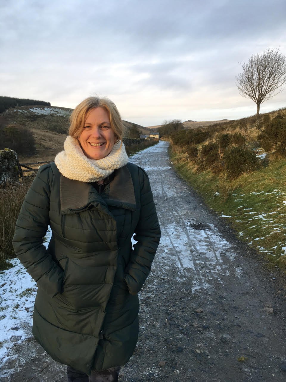 A woman in a dark green winter coat and a cream-colored scarf standing on a snow-dusted dirt path in a rural landscape with rolling hills and a leafless tree against a cloudy sky.