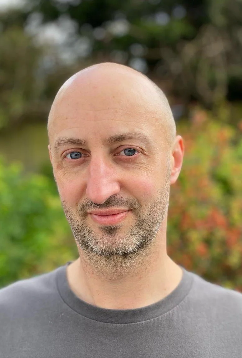 A man with a bald head wearing a grey top against a backdrop of red flowers and a green bush