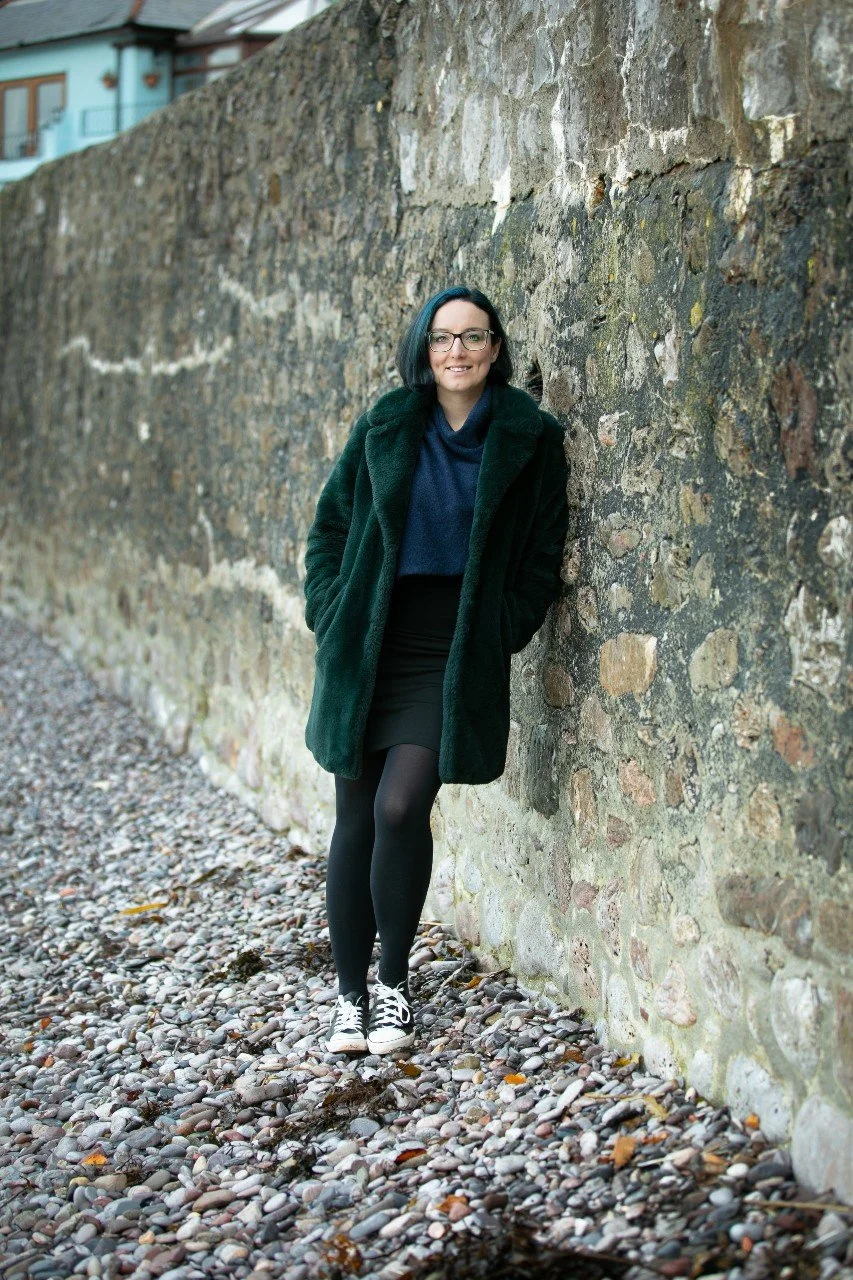 A woman with black hair, glasses, a dark green coat, black skirt, black tights, and trainers standing against a stone wall on a pebble-covered ground.