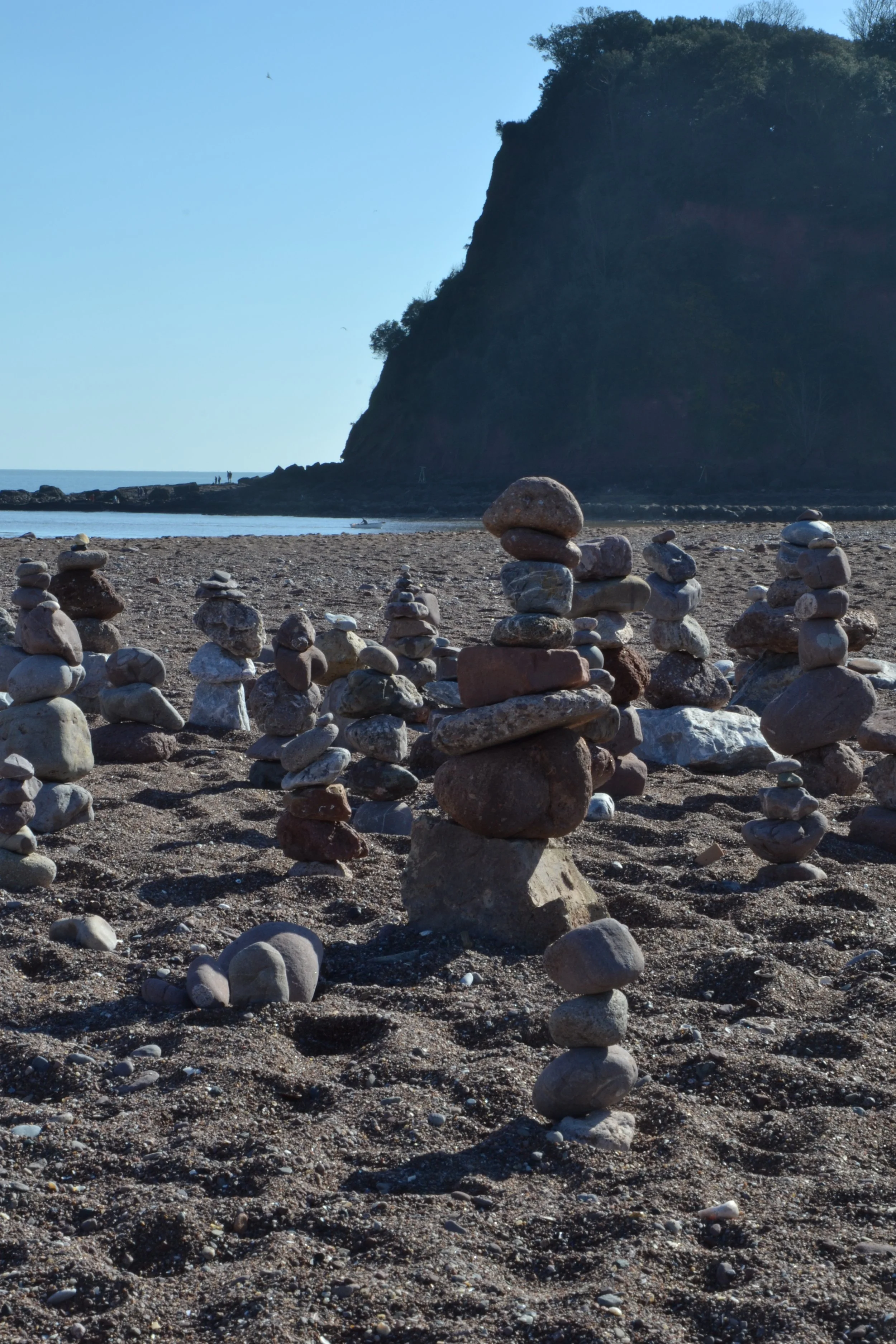 A beach with numerous small stacked rocks in the foreground, a cliff covered with trees on the right, and a calm ocean in the background under a clear blue sky.