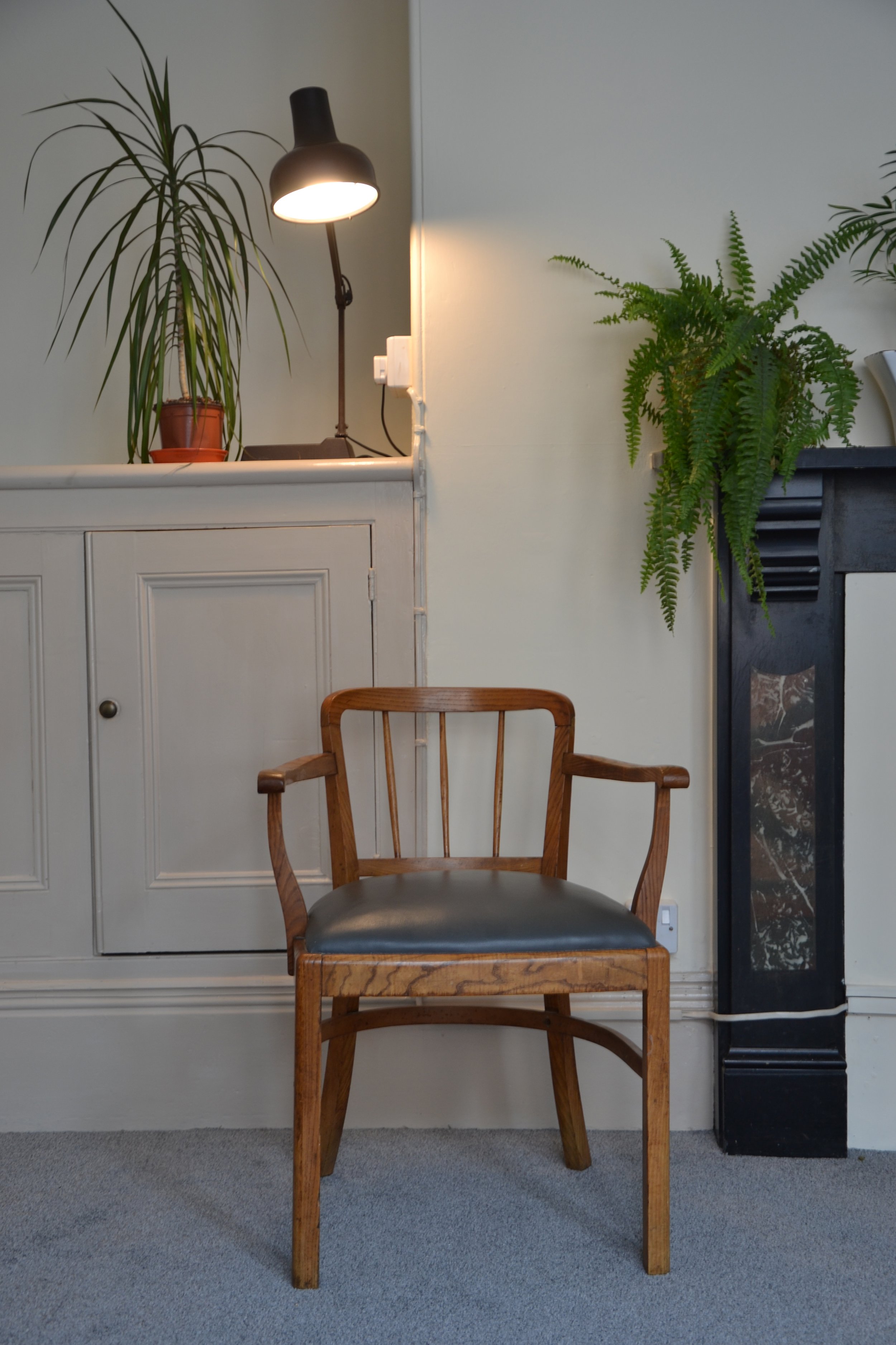 A wooden chair with a padded black seat positioned in front of a built-in cabinet and fireplace mantel. On the mantel, there are green plants and a black desk lamp turned on.