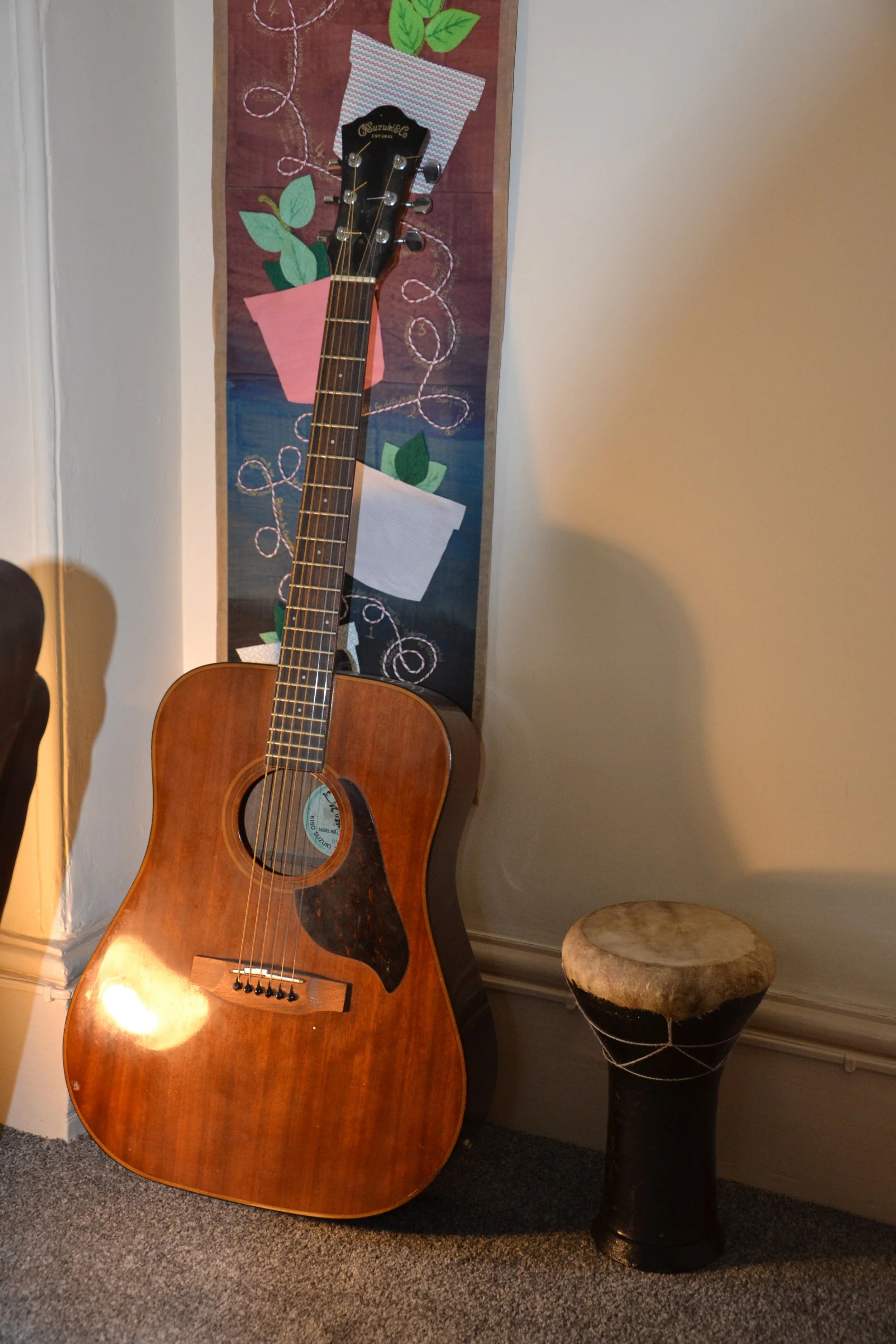 An acoustic guitar leaning against a wall next to a small drum. There is a colourful fabric wall hanging with plant and vase designs behind the guitar.