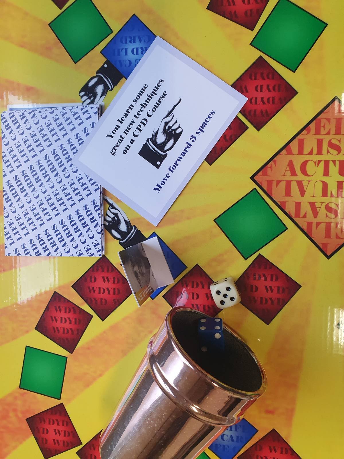 Board game with red and green squares, cards, a small photo, and a die. One card states learning new techniques on a CPD course, and the board has spaces labeled with What would you do?