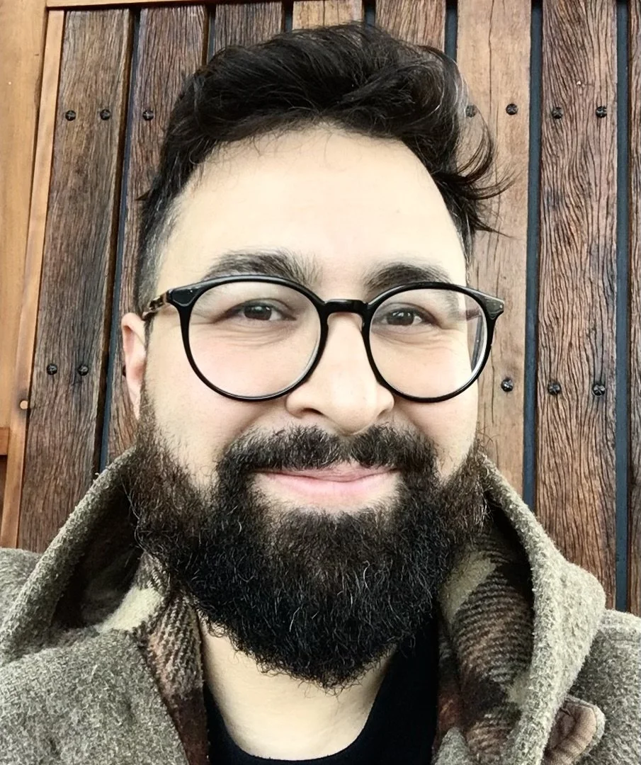 Sami Choudhury (tutor) with glasses, a beard, and short dark hair, wearing a brown coat, sitting in front of a wooden wall.