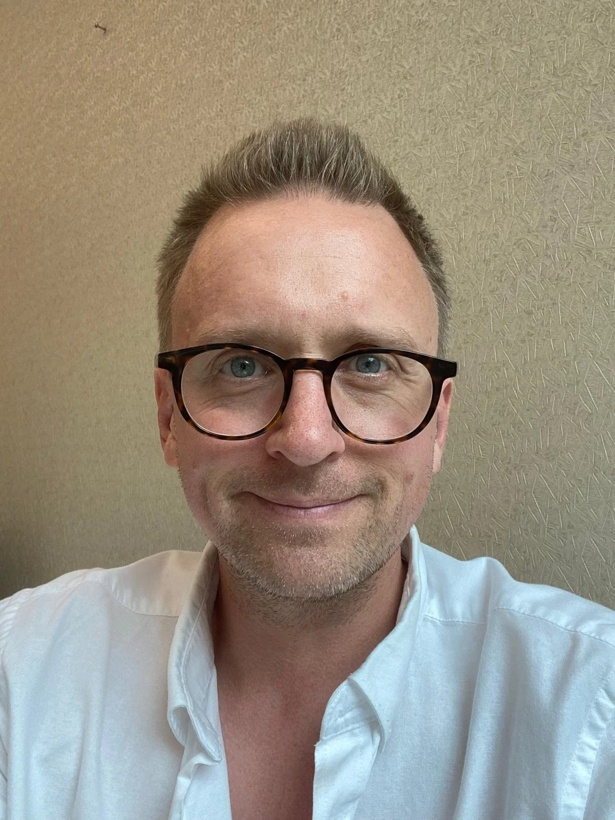 Patrick Carter (Training Co-ordinator) with light skin and glasses smiling at the camera, wearing a white shirt, against a beige textured wall.