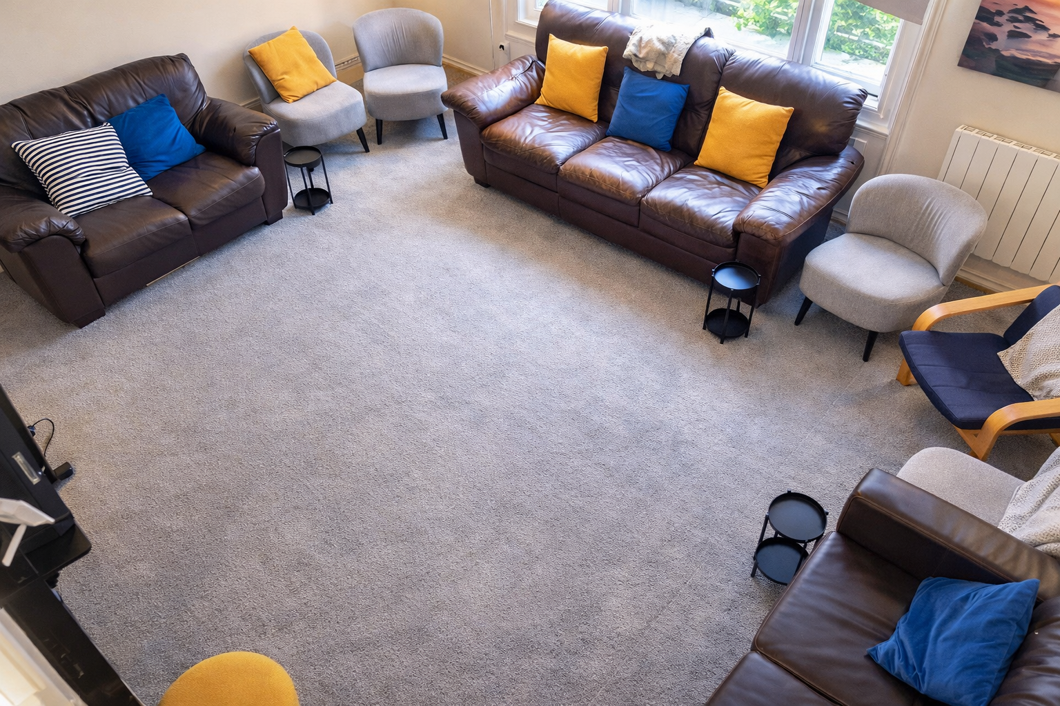 Living room with three sofas and eight cushions facing each other, two on the left and six on the right, small black tables, and two grey armchairs. Large windows with greenery outside, carpeted floor, and a painting on the wall.