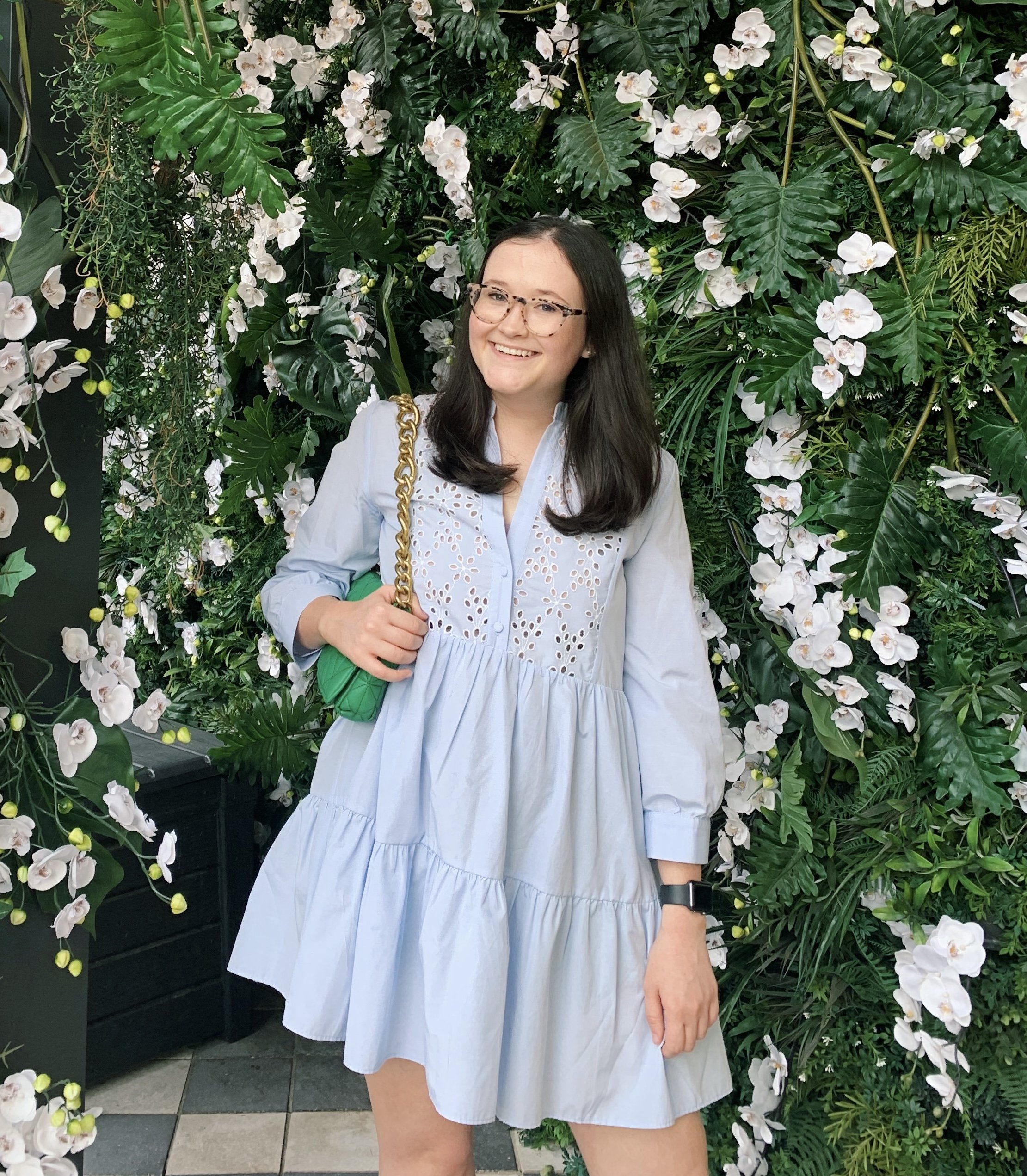 A woman with long dark hair and glasses smiling, standing in front of a lush green wall with white orchids and tropical leaves, dressed in a light blue dress and carrying a green quilted purse with a gold chain strap.