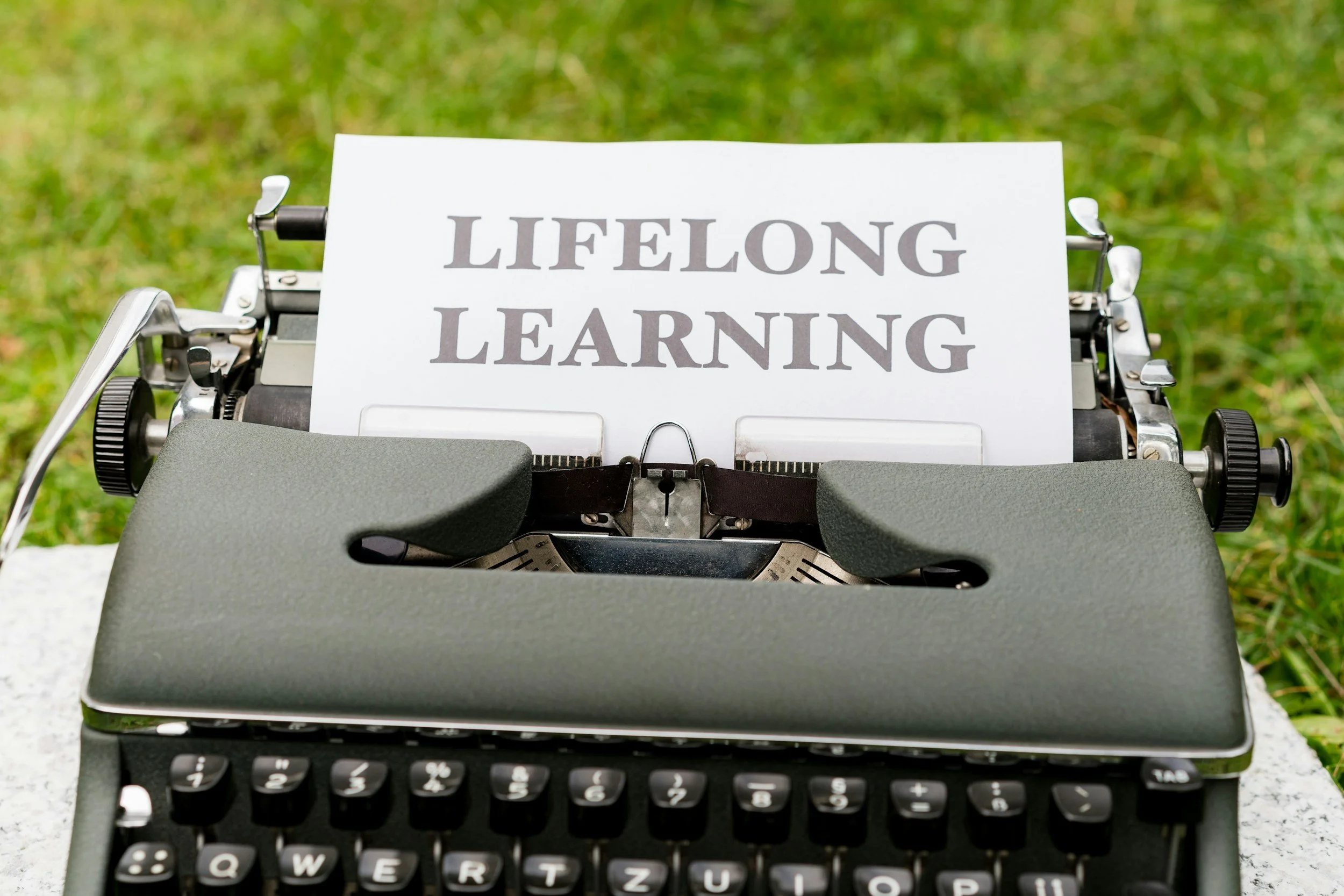 Eine Schreibmaschine mit einem Zettel, auf dem 'LIFELONG LEARNING' steht, vor einem grünen Rasen im Hintergrund.