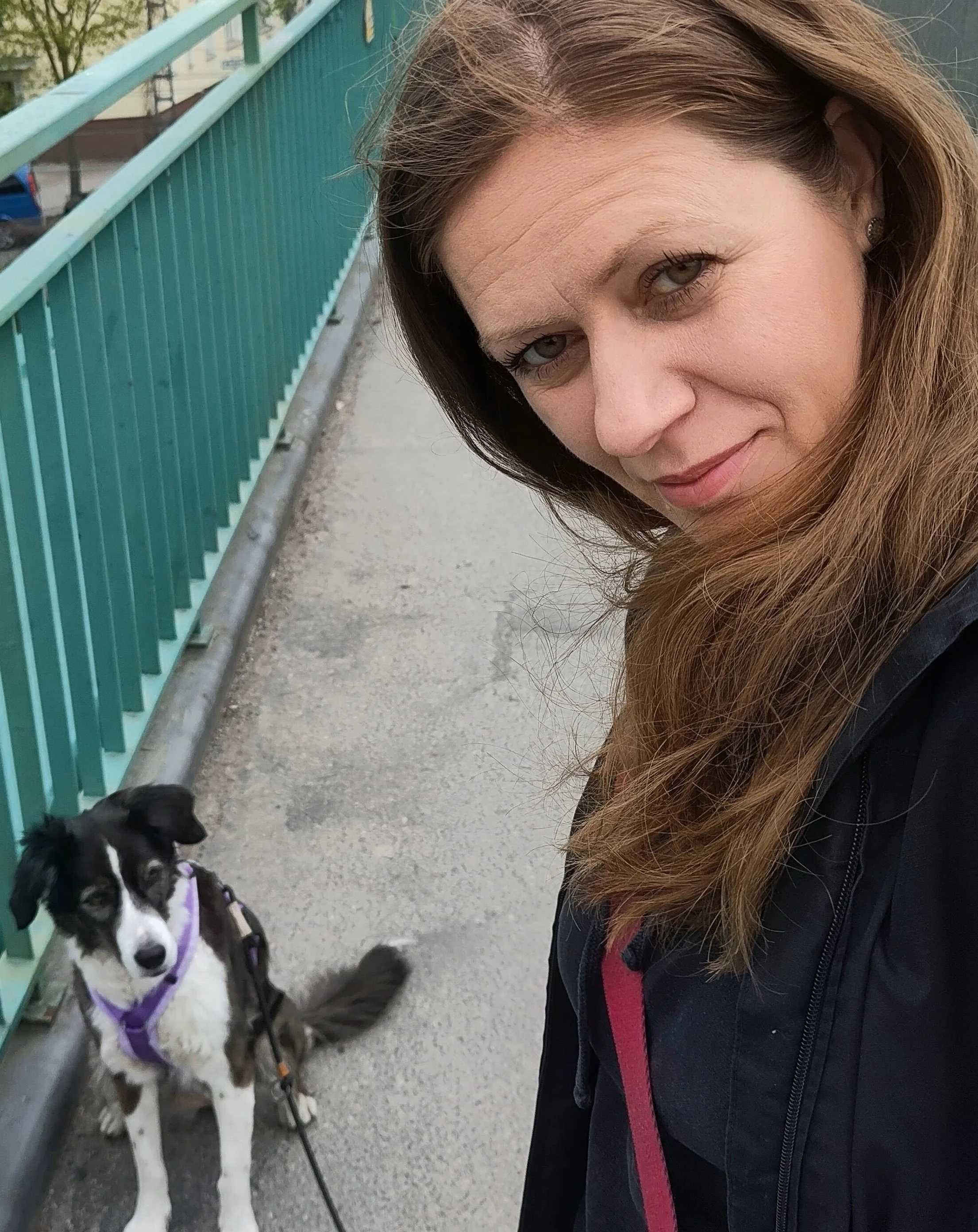 Eine Frau mit braunen, lockigen Haaren posiert neben einem schwarz-weißen Hund auf einem Gehweg. Im Hintergrund befindet sich eine grüne Metallbrüstung und es sind Bäume und Gebäude sichtbar.