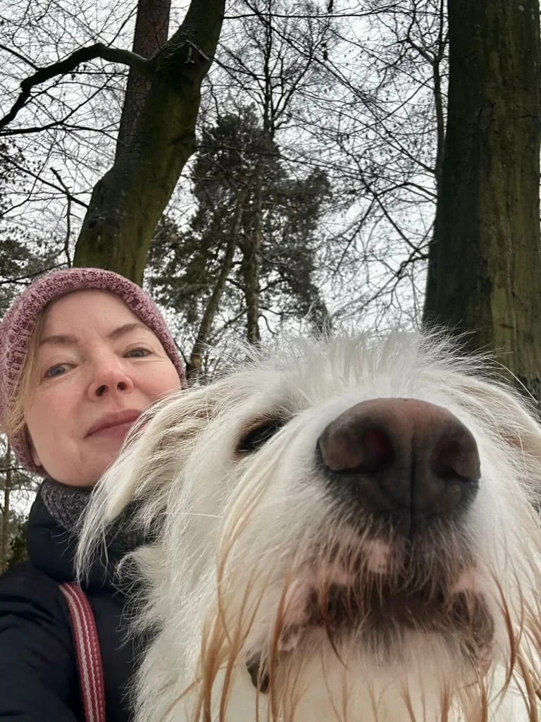 Frau im Wald mit pinkem Hut und schwarzen Jacke, Nahaufnahme eines weißen, schmuddeligen Hundes mit nassem Gesicht, Bäume im Hintergrund