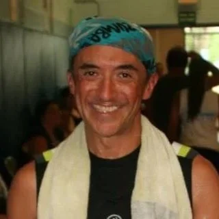 Smiling man wearing a blue swim cap and black sleeveless shirt, indoors with people in the background.