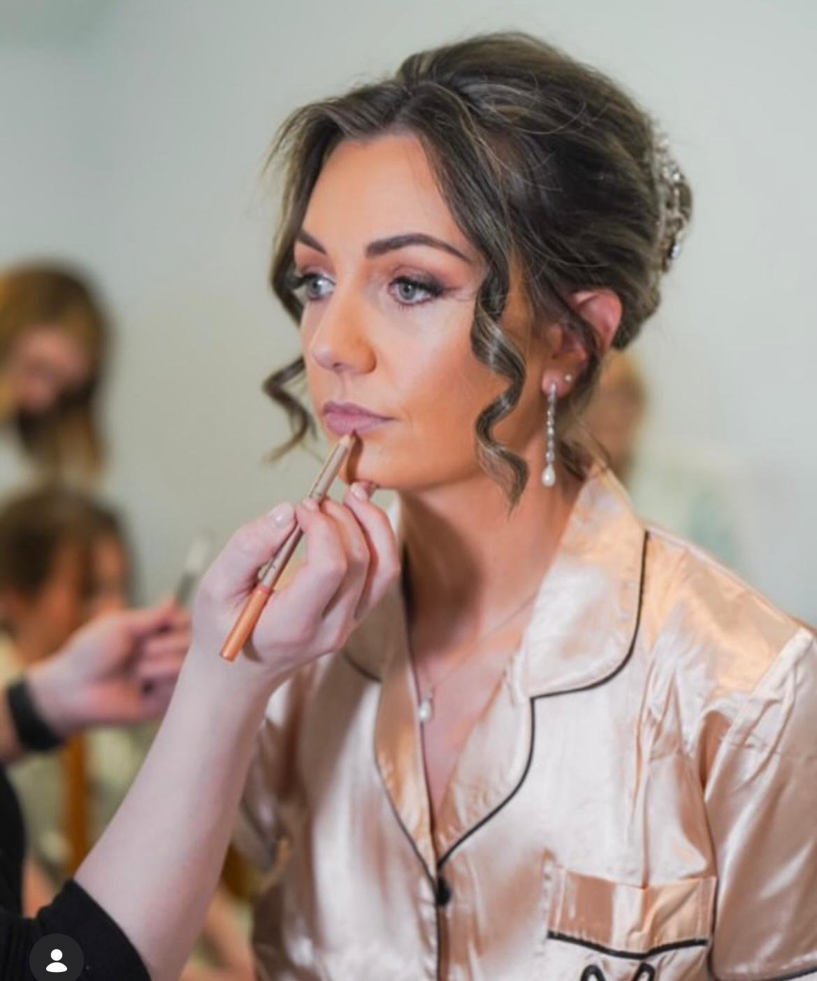 Woman with styled hair and pearl earrings getting makeup applied on her lips.