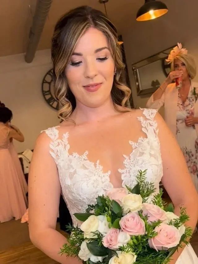 A bride in a white wedding dress holding a bouquet of pink and white roses, smiling softly at a wedding reception.