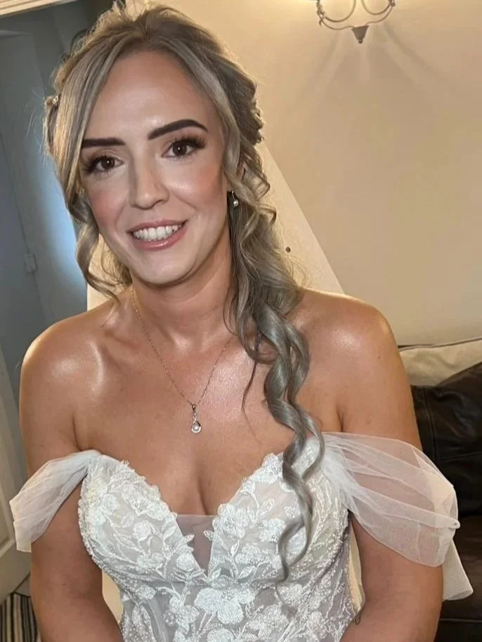 A woman with blonde hair styled in loose curls with a grey tint, wearing a white lace off-the-shoulder dress and a necklace, smiling at the camera.
