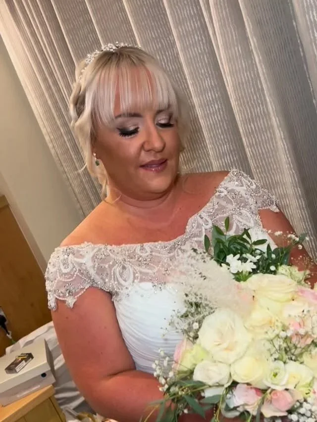 A woman in a wedding dress holding a bouquet of white and pink flowers, standing in front of a curtain.