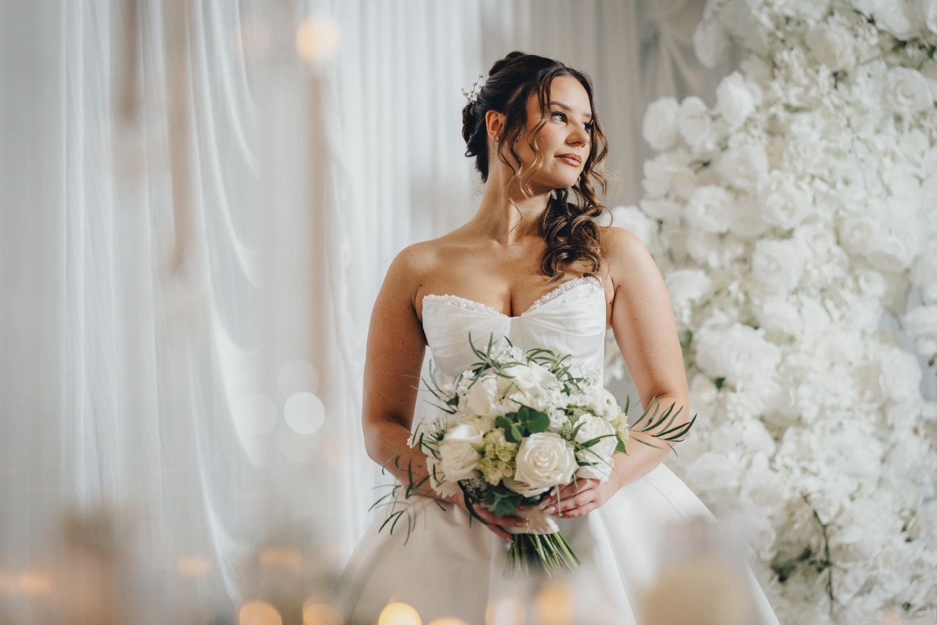 A bride in a white wedding dress holding a bouquet of white roses and greenery, standing in front of a white floral backdrop on her wedding day.