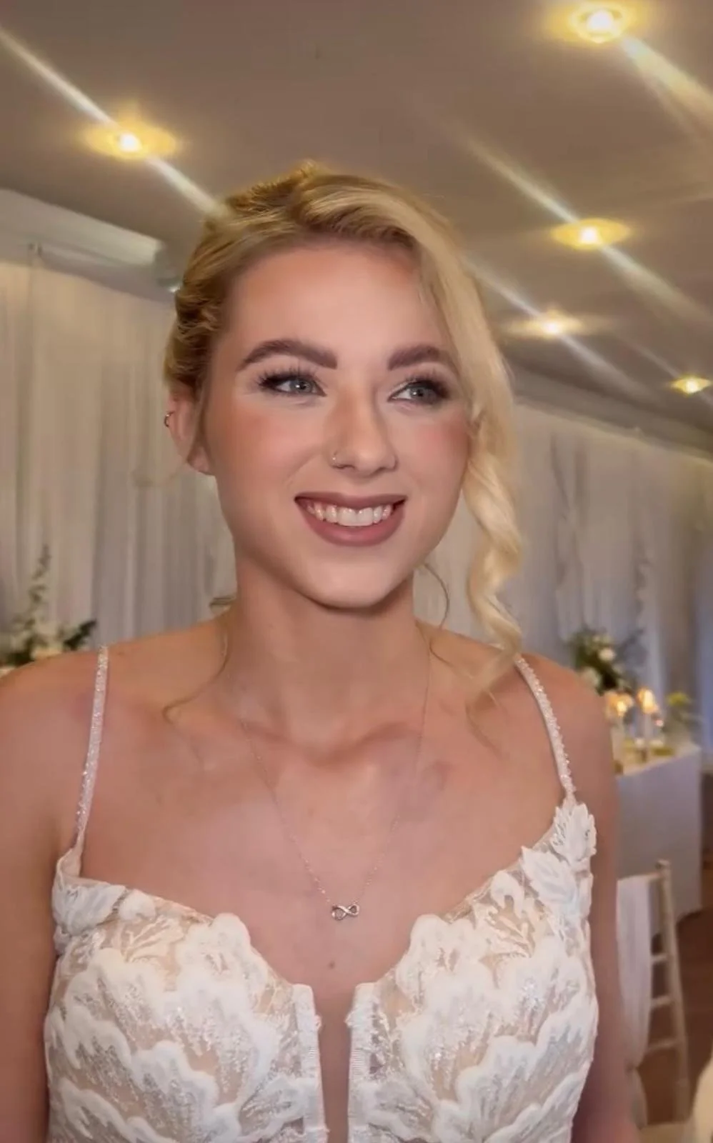A smiling woman with blonde hair styled in loose curls, wearing a white lace dress with thin straps and a small infinity necklace, standing in a decorated indoor setting.