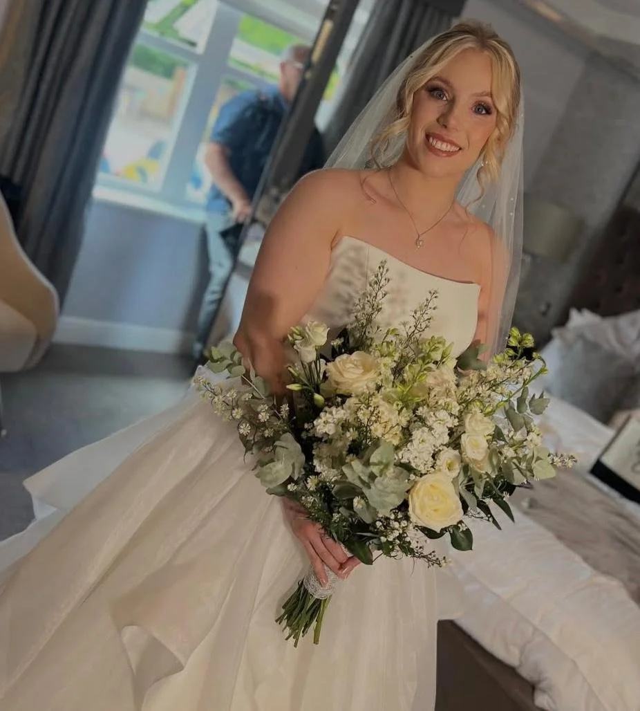 Bride in wedding dress holding a large bouquet of white and green flowers, smiling.