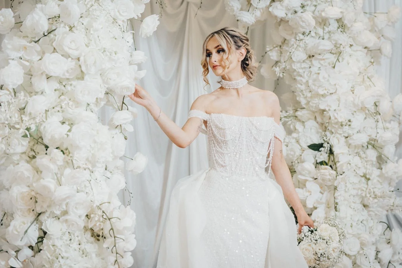 A woman in a wedding dress standing among white floral arrangements, holding a bouquet, in a bridal setting.