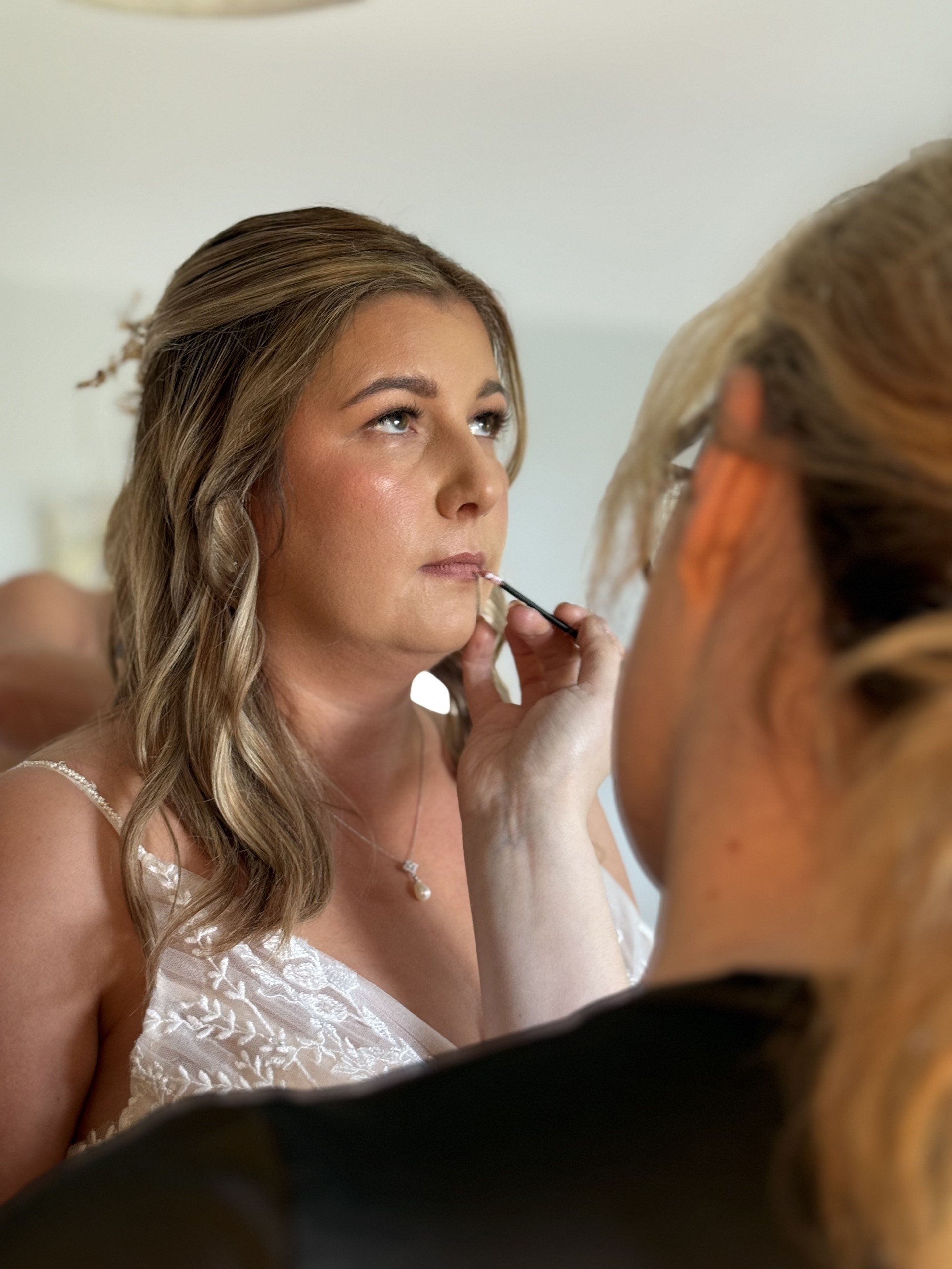 A woman getting her makeup applied by a makeup artist. The woman has light brown, wavy hair and is wearing a white dress with embroidery and a pearl necklace. The makeup artist's face is not visible.