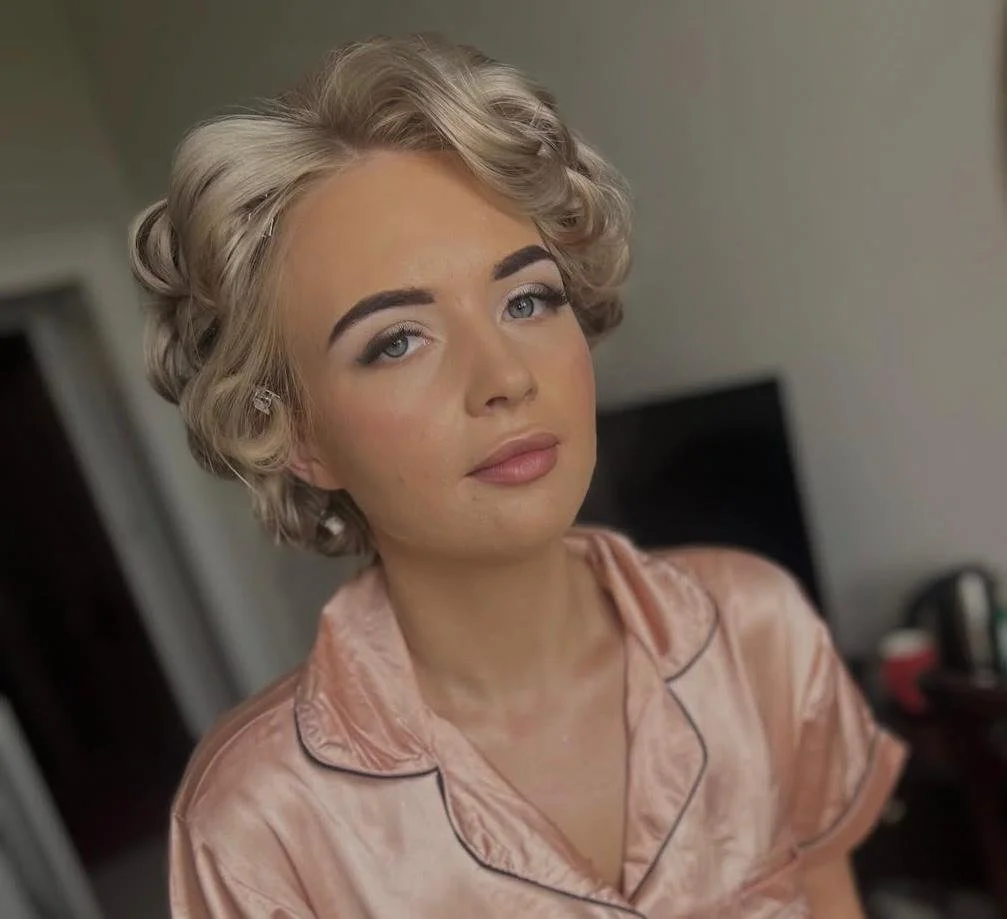 Close-up of a young woman with blonde, curled hair wearing a peach satin pajama top, looking at the camera.
