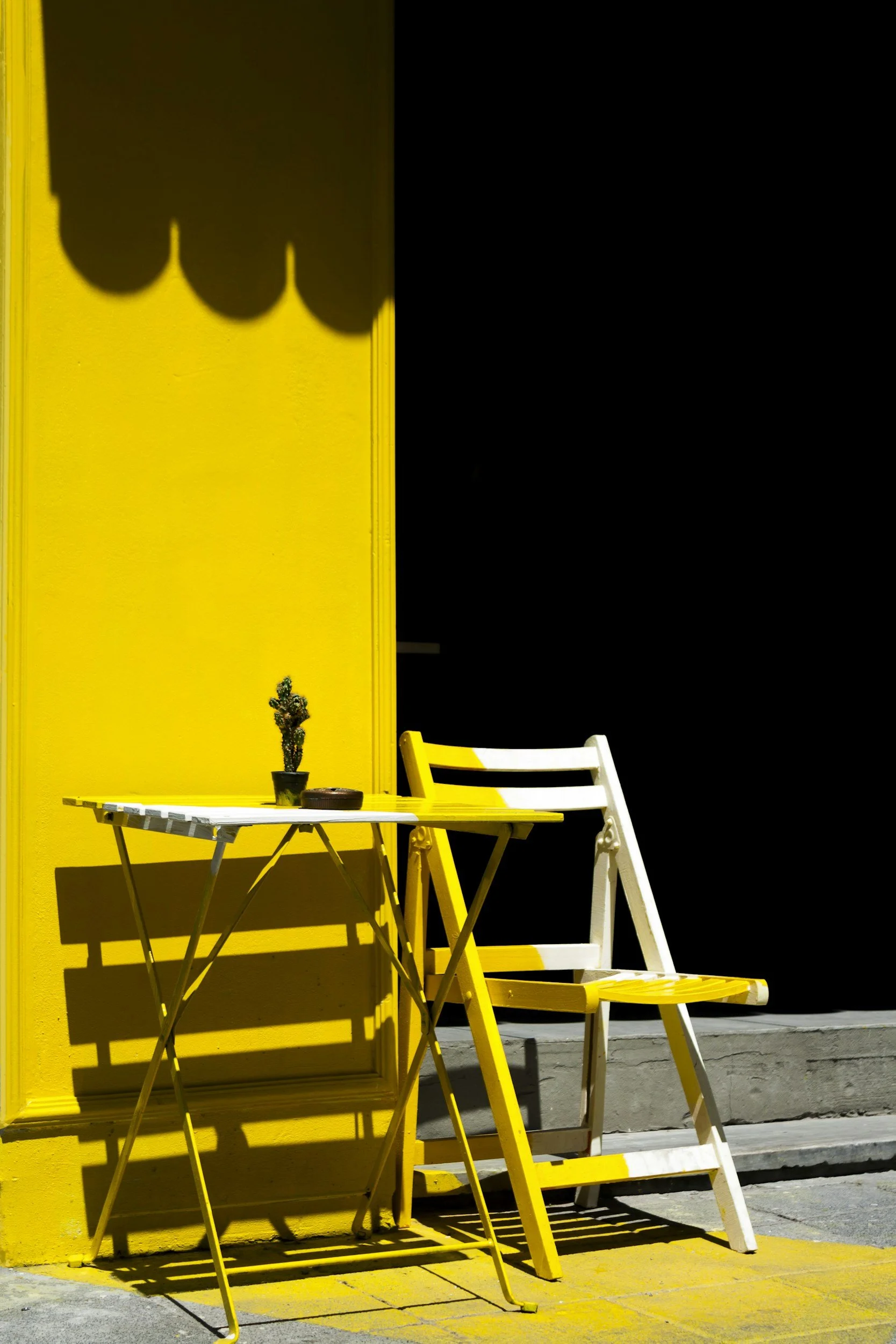 Yellow wall with a matching yellow table, white & yellow chair, small potted plant, shadow of a canopy, and a black dark background.
