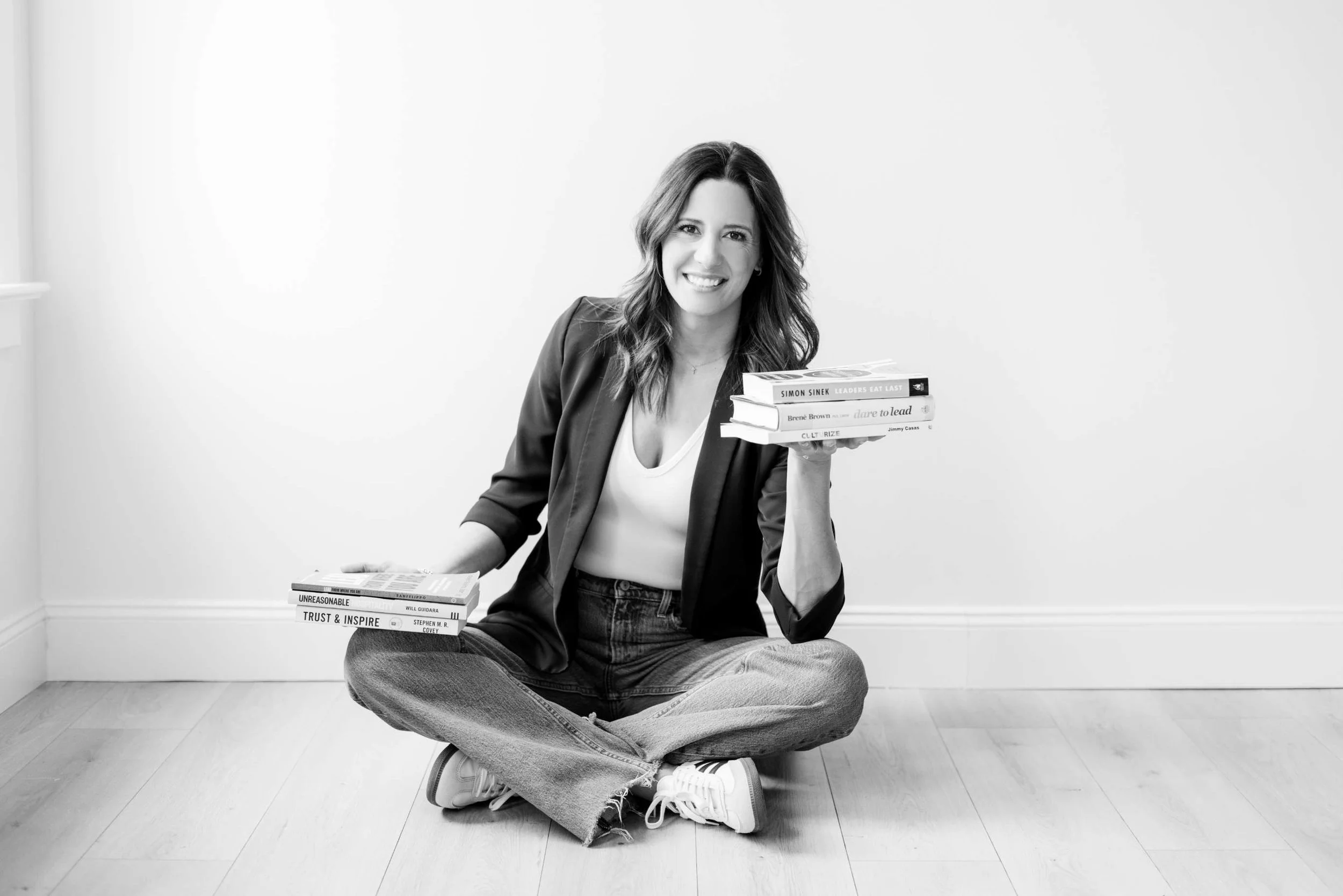 A woman with shoulder-length hair sitting on the floor, cross-legged, wearing a blazer, jeans, and sneakers, holding a stack of books with titles visible, in a plain room with a white wall.