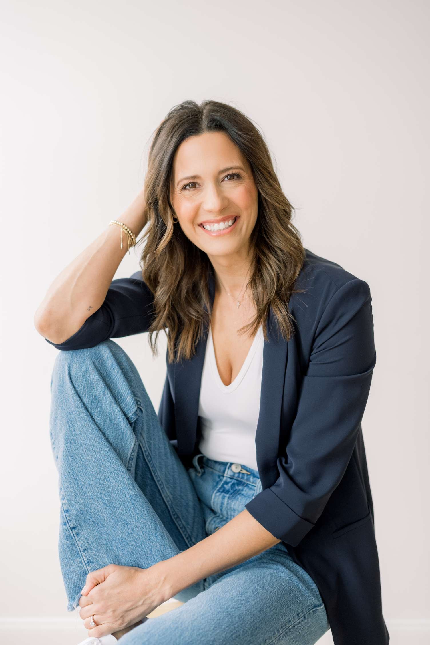 A woman with brown wavy hair, smiling, wearing a dark blazer over a white top, sitting with her legs crossed, and a woman with straight brown hair, wearing a black top and blue jeans, sitting on her lap with her arms around her neck, in front of a plain white wall.